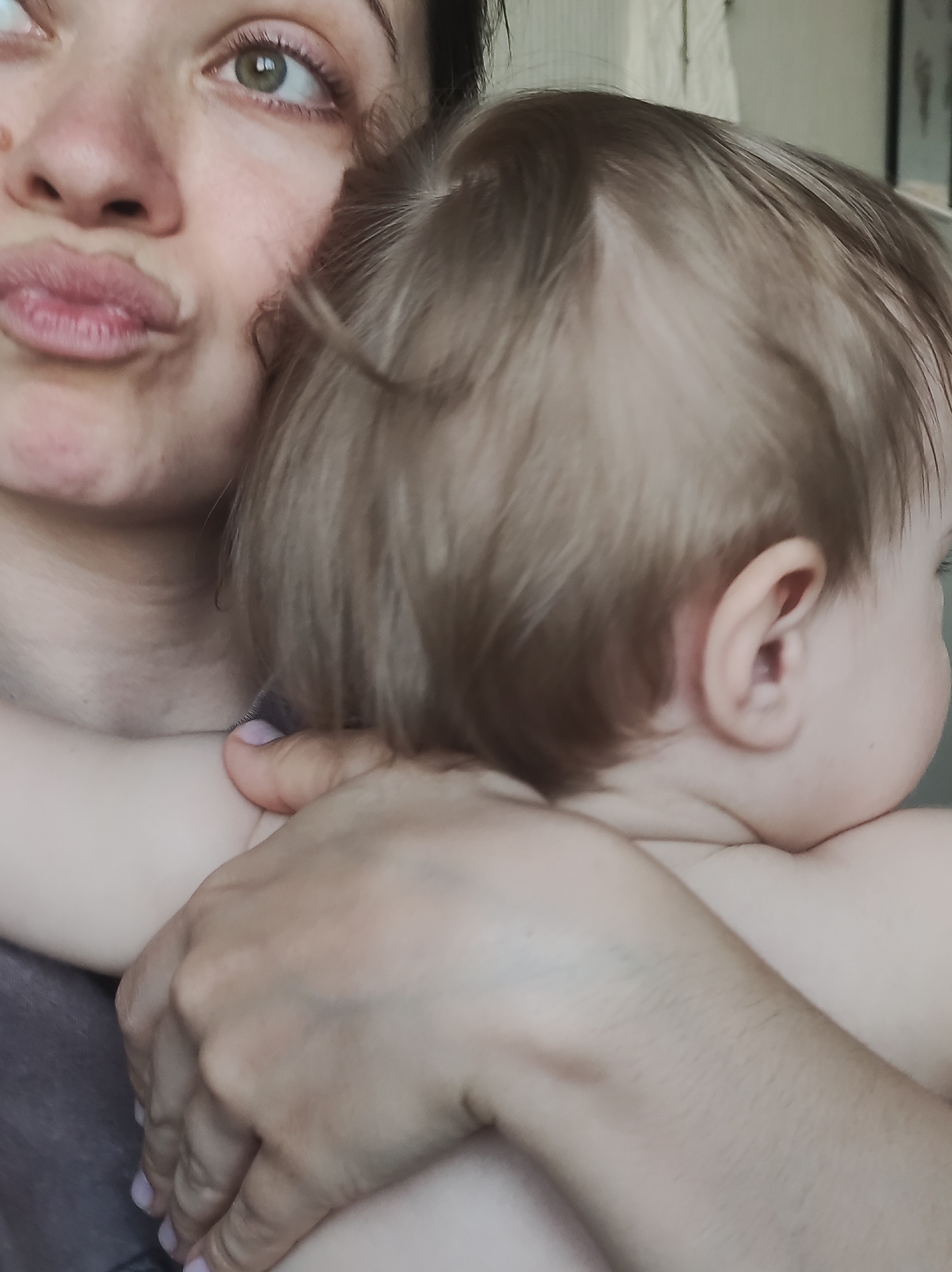 This intimate, close-up indoor photograph captures a tender moment between a woman and a baby. The scene is tightly framed, focusing on their intertwined forms, suggesting a personal and loving connection, likely within a home setting in Chaikovsky, Russia.
The woman occupies the left side of the frame, characterized by fair skin, striking green or hazel eyes, and partially visible dark hair. Her lips are pursed in a "kissing face" or playful expression, conveying affection. Her skin exhibits natural texture, and her visible fingernails are neatly manicured with a pale, possibly lavender, polish. One of her hands is seen securely holding the baby, with fingers gently resting on the baby's back or shoulder. Her forearm is also visible, showing fine, light hair and a faint bluish vein or mark.
The baby, held close to the woman's body, is positioned on the right side of the frame with its back to the camera. Its head is turned slightly to the right, resting against the woman's shoulder or chest. The baby has soft, light-colored (blonde to light brown) fine hair, which shows natural variations in density on the scalp, common in infants. Its skin is fair and smooth, and a small, delicate ear is clearly visible. The baby appears to be unclothed or wearing minimal clothing on its upper body.
The background is softly blurred, indicating a shallow depth of field that keeps the focus entirely on the subjects. It appears to be an indoor setting with light-colored walls or curtains. The lighting seems natural, suggesting it is daytime. No explicit text is visible in the image. The overall impression is one of warmth, parental love, and a quiet, cherished moment of bonding.