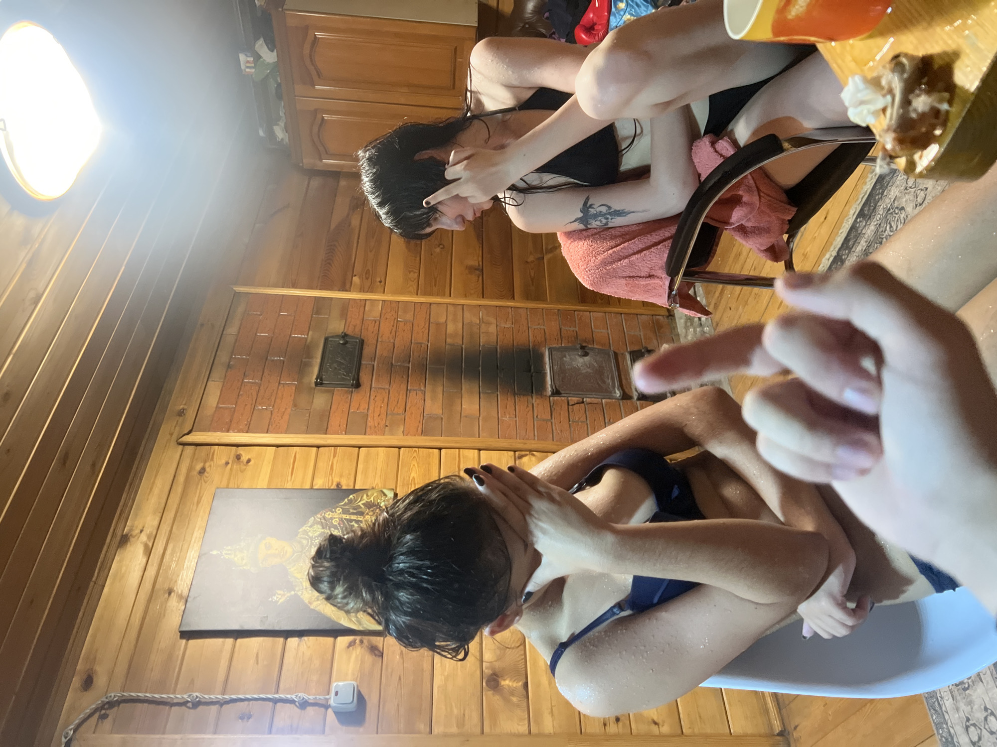 The image depicts two young women in a wooden-paneled room, possibly a sauna or a similar recreational space. The room features exposed wooden beams on the ceiling and brickwork on one wall, where a black metal insert is visible. A framed artwork depicting a person in ornate attire hangs on the wooden wall. One woman, on the left, is wearing a dark blue bikini top and has her face covered with her hands. Water droplets are visible on her skin. The other woman, on the right, wears a black bikini top and is seated on a black chair draped with a pink towel. She has a tattoo on her left arm and is making a rude gesture with her right hand. A cup and a small plate with food are on a table in the foreground. The scene is lit by an overhead light fixture. The image is taken from a low angle, with a hand in the foreground obscuring part of the frame. The location is identified as Solontsy, Russia.