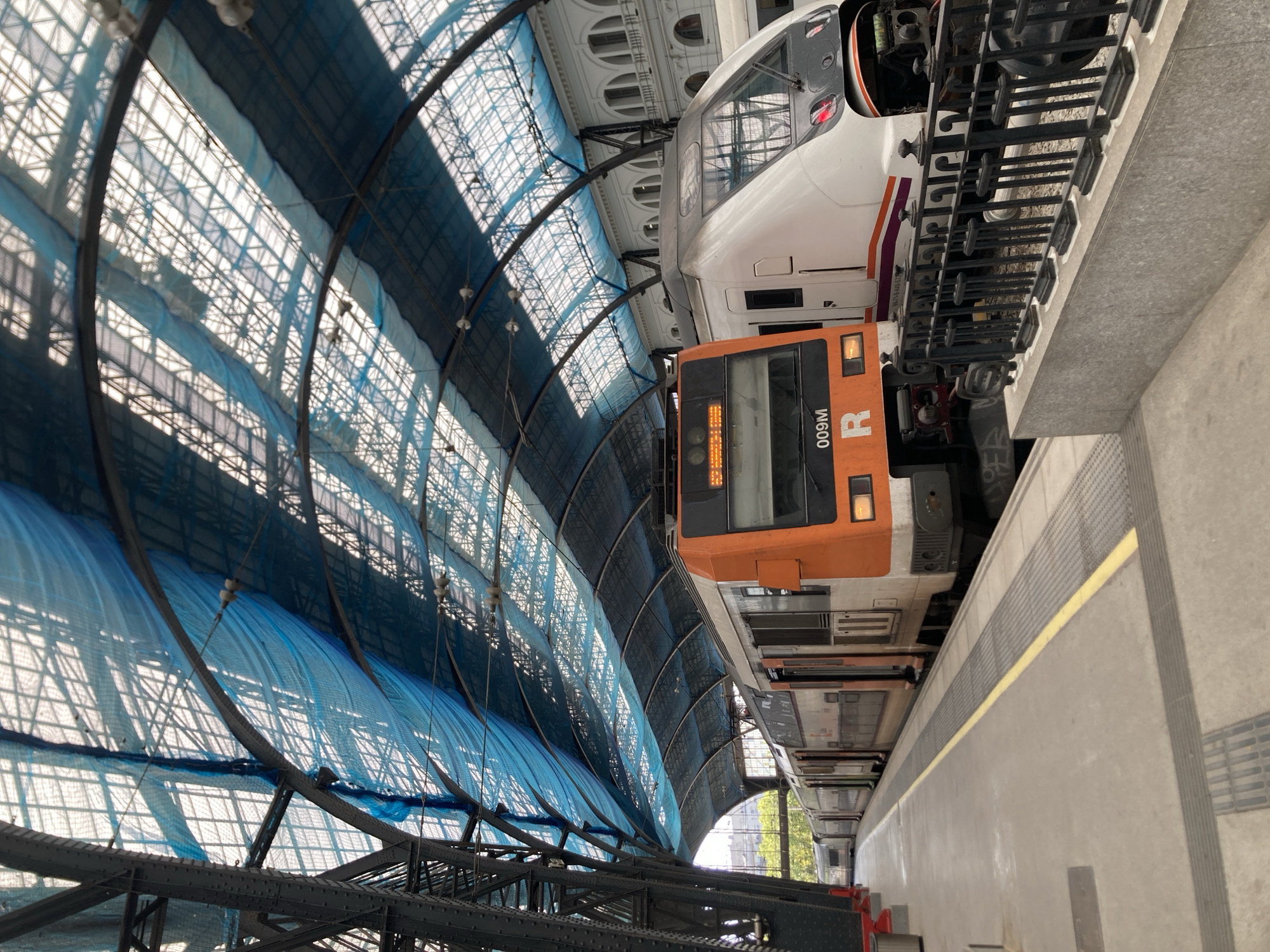 The image shows two trains at a station in Barcelona, Spain. The train on the left is orange and white, and the train on the right is white and purple. Both trains are on parallel tracks, and there is a platform between them. The station has a distinctive arched roof with a blue net covering. The architectural style of the buildings in the background suggests a historic European train station. The scene appears to be captured from the platform, looking towards the trains and the station's roof structure. The lighting is bright, indicating daytime.