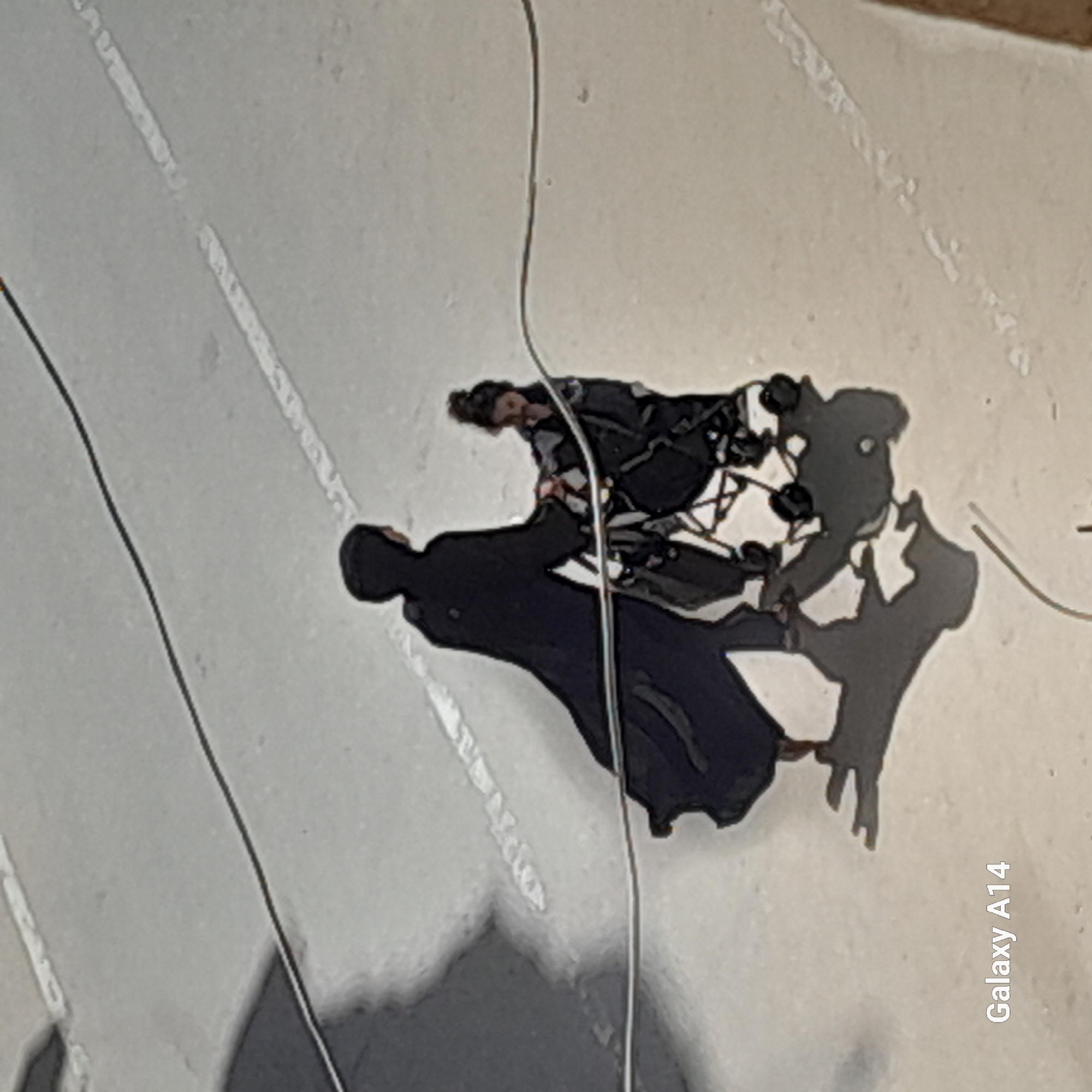 A high-angle shot shows a person in a wheelchair being pushed by another person wearing a dark robe or cloak on a light-colored surface, possibly pavement, with white dashed lines indicating a lane. There's a thin wire or string visible in the frame. The location appears to be an unidentified outdoor area. The person in the wheelchair seems to be interacting with the person pushing the wheelchair. The environment consists of paved surfaces with lane markings. The smartphone model "Galaxy A14" is visible in the lower right corner, likely indicating the device used to capture the image.