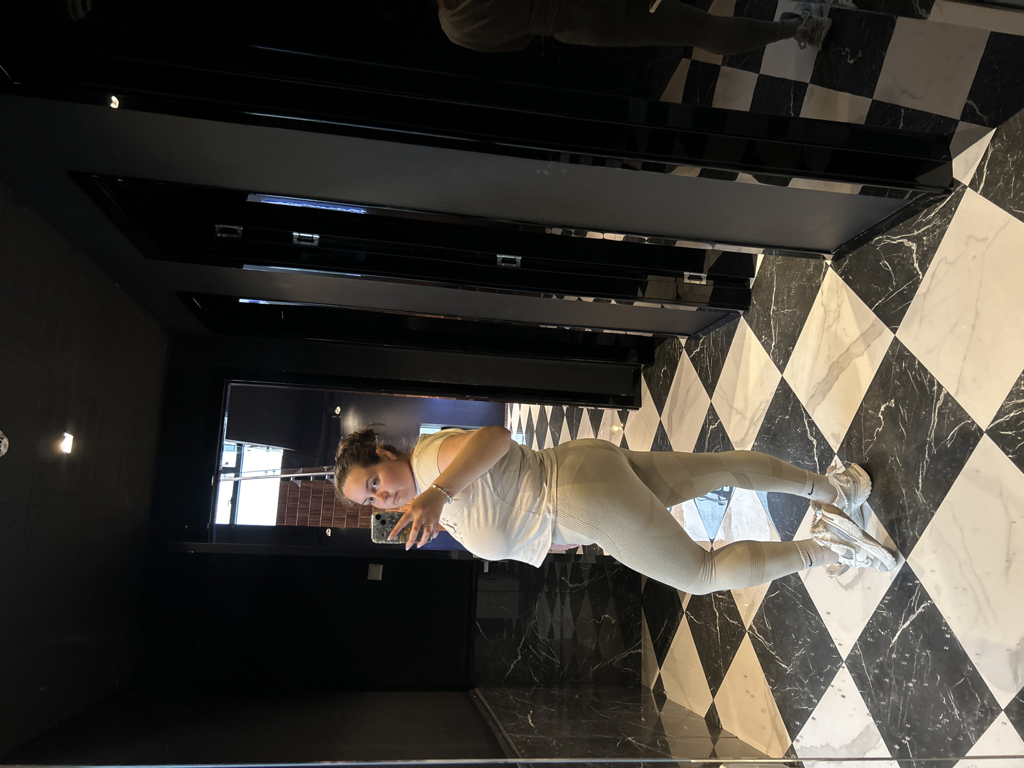 A woman is taking a selfie in what appears to be a bathroom or fitting room with black and white checkered floors and black glossy walls. She is wearing a light-colored athletic outfit and sneakers. The room features dark stall doors and a small window reflecting part of a building. The location is likely in Plovdiv, Bulgaria, based on the prompt, but there's no specific architectural or contextual detail in the image to confirm this.