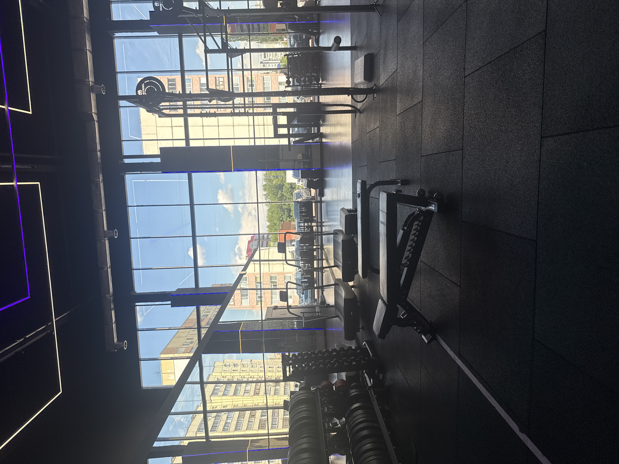 The image shows the interior of a modern gym with large windows looking out onto a city street. The gym is equipped with various exercise machines, including treadmills and weightlifting equipment. There are also weight racks filled with plates and dumbbells, as well as adjustable benches. The flooring is dark, textured rubber, and a white line is visible on the floor. The gym features contemporary lighting, with neon purple and white accents on the ceiling. Outside the windows, apartment buildings and trees are visible, along with parked cars. The scene is brightly lit by natural daylight filtering through the windows.