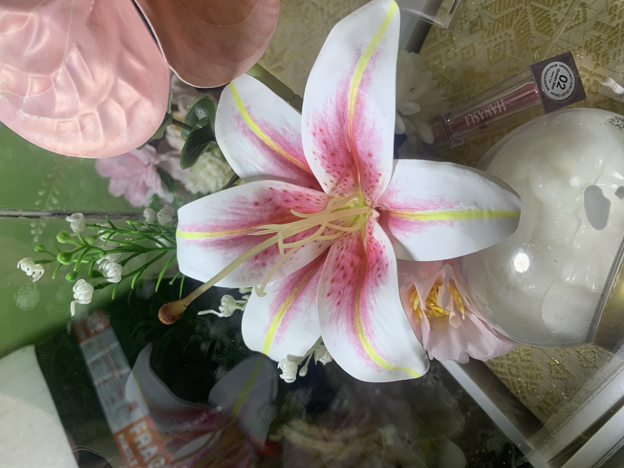 The image is a close-up of a floral arrangement, featuring a prominent white and pink lily with yellow accents. Surrounding the lily are smaller white flowers and green foliage. In the background, a soft pink anthurium flower is visible. A clear plastic container with a light yellow, patterned insert is present at the bottom, containing a small, pink lipstick tube labeled "HANASUI" and "QUEEN BEE." The reflection in the glass shows an orange and white "FRAGILE" sticker. The scene appears to be indoors, possibly a display or gift arrangement. There are no people or specific landmarks visible that would indicate a location in Kuala Pembuang, Indonesia.