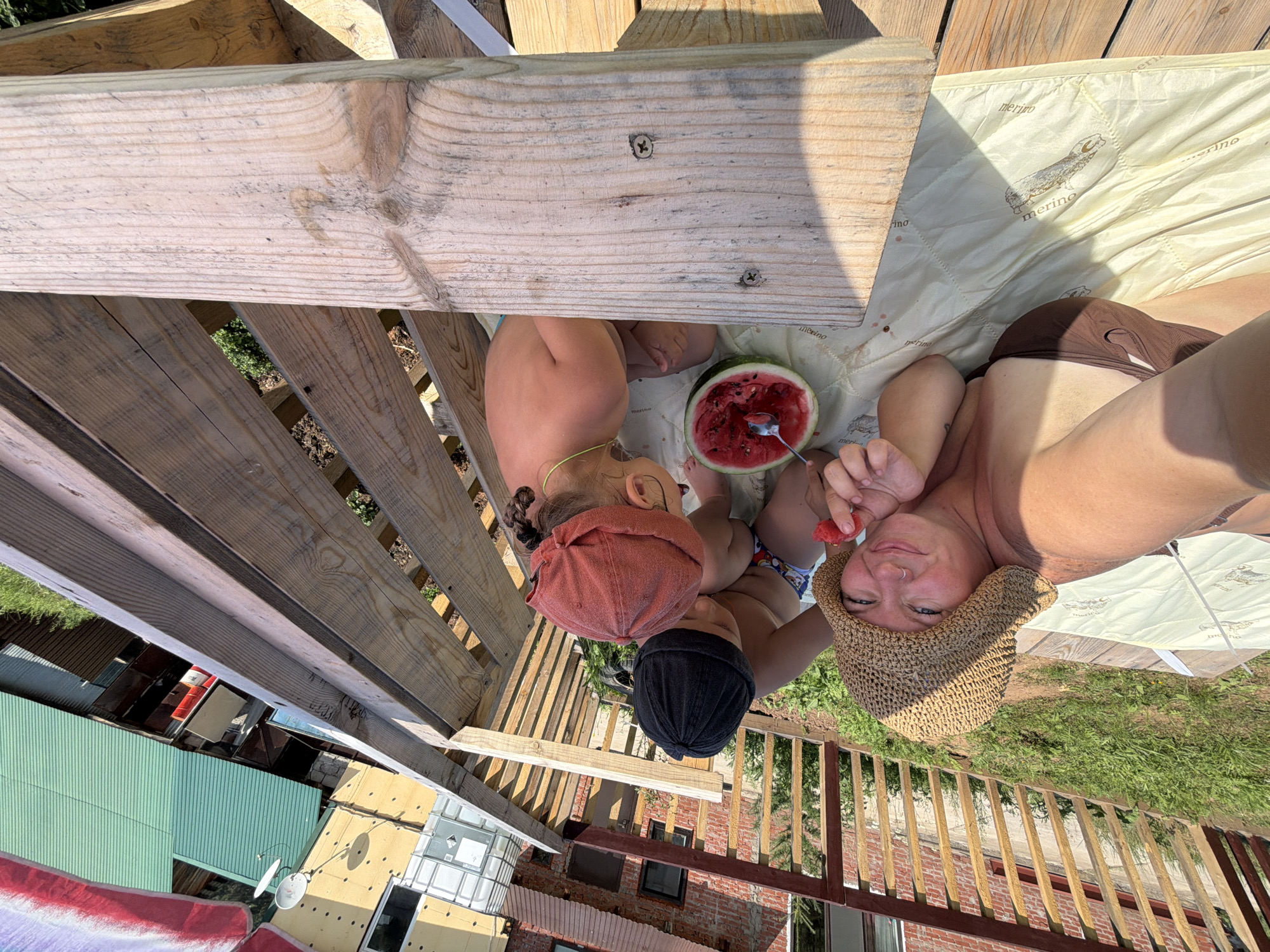 In Chaikovsky, Russia, a woman and two young children are on a wooden deck on a sunny day. The woman, wearing a straw hat and a brown swimsuit, is lying down and holding a piece of watermelon. To her right, a child in a black beanie is seated and looking down at the watermelon. Beside that child, another child in a red beanie is also seated and looking at the watermelon. A half-eaten watermelon with a spoon in it sits on a light-colored mat between them. In the background, there is a wooden fence and buildings with colorful corrugated metal roofs. The scene suggests a casual outdoor gathering, likely in a backyard or recreational area.
