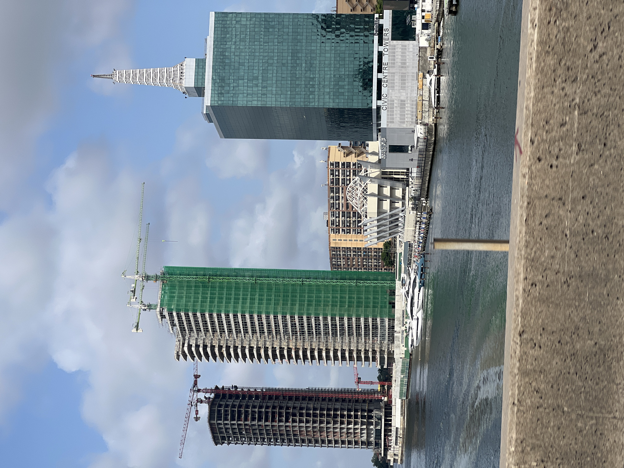 The image shows a waterfront view in Ikoyi, Nigeria. From left to right, three high-rise buildings dominate the skyline. The leftmost building is under construction with visible scaffolding and a red construction crane. The middle building is also under construction, draped in green netting, and has a green construction crane. The rightmost building appears to be completed, featuring reflective glass panels and the words "CIVIC CENTRE TOWERS Deloitte" vertically aligned on its side. A body of water separates the viewer from the buildings. Numerous boats are docked along the waterfront. A concrete barrier is in the foreground. The sky is blue with scattered clouds.