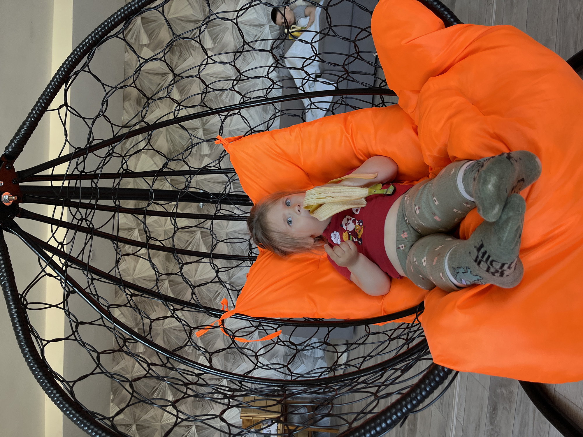 A young child is lying in an orange-cushioned, black wire hanging chair. The child is fair-skinned with blue eyes, wearing a burgundy t-shirt with cartoon characters, patterned green leggings, and green socks. The child appears to be eating a peeled banana.
In the background, through the wires of the hanging chair, another child is lying on a gray couch with a white blanket, also eating a banana. The background indicates an indoor setting with patterned wallpaper or wall covering. The image was taken in Chaikovsky, Russia.