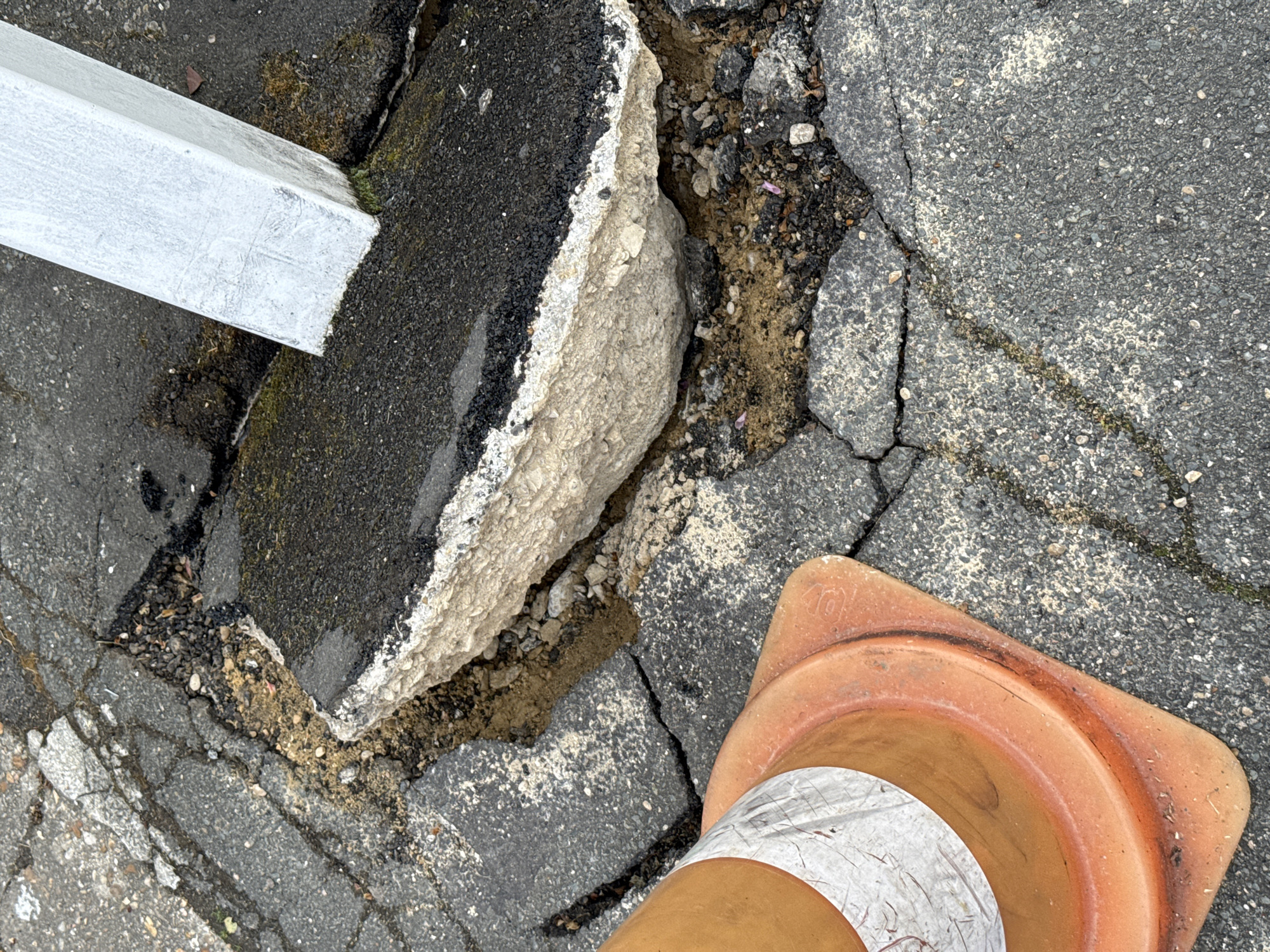 The image depicts damaged asphalt pavement in Domont, France, near a white painted post. A chunk of asphalt is broken away, revealing a layered composition of asphalt and concrete. Cracks traverse the surrounding pavement. A traffic cone with a white strip is placed near the damage.