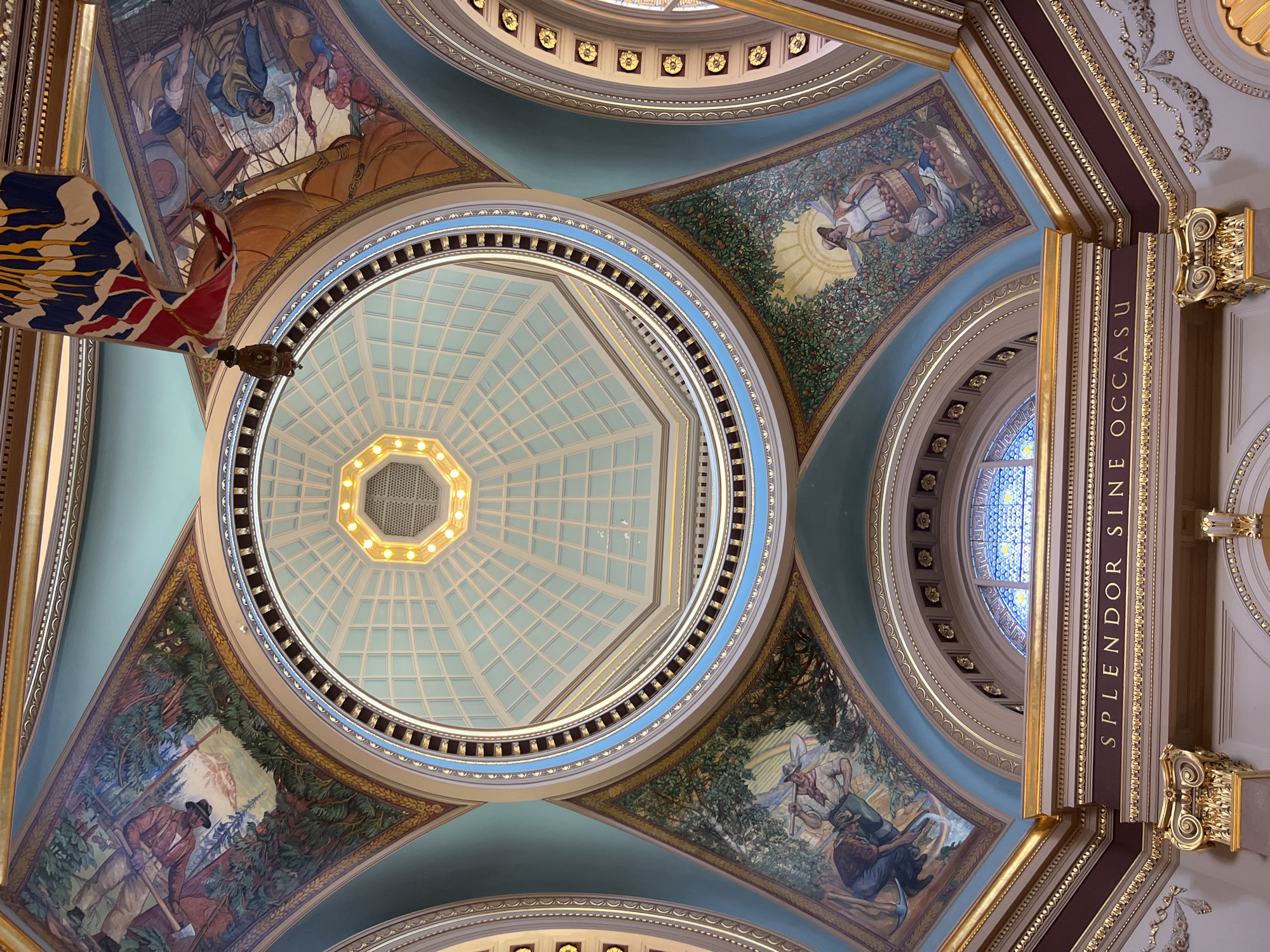 The image shows the interior of the dome of the Legislative Assembly of British Columbia in Victoria, Canada. The dome is octagonal and features a skylight at its apex, which is illuminated. The interior is richly decorated with murals, gold accents, and a central inscription.
The murals depict scenes of early British Columbia, including images of people working in forests and harvesting fruit. These murals are set within arched sections of the dome.
The inscription on a frieze reads "SPLENDOR SINE OCCASU," which is Latin for "Splendor Without Setting." The frieze is above a stained-glass window.
A Canadian flag, partially obscured, hangs from the ceiling in the upper right portion of the image. The overall impression is one of grandeur and historical significance.