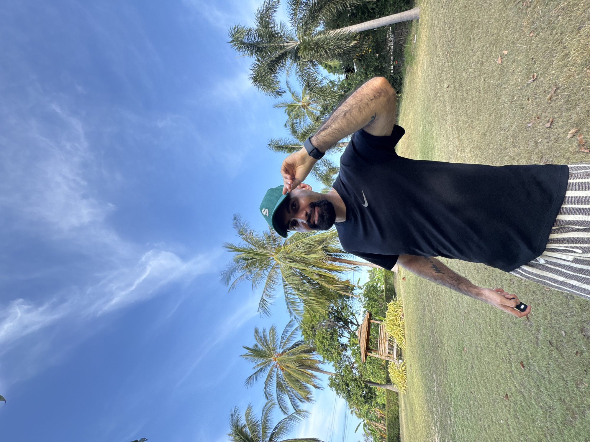 In the city of Ko Samui District, Thailand, a man with dark hair and a beard is posing for a photo outdoors. He is wearing a black t-shirt with a Nike logo and a green baseball cap with an "S" logo. He is holding the brim of his cap with his right hand. The background shows a grassy lawn, palm trees, and a clear blue sky with wispy white clouds. There is a wooden gazebo with a thatched roof in the middle ground. The man is holding a small black object in his left hand.