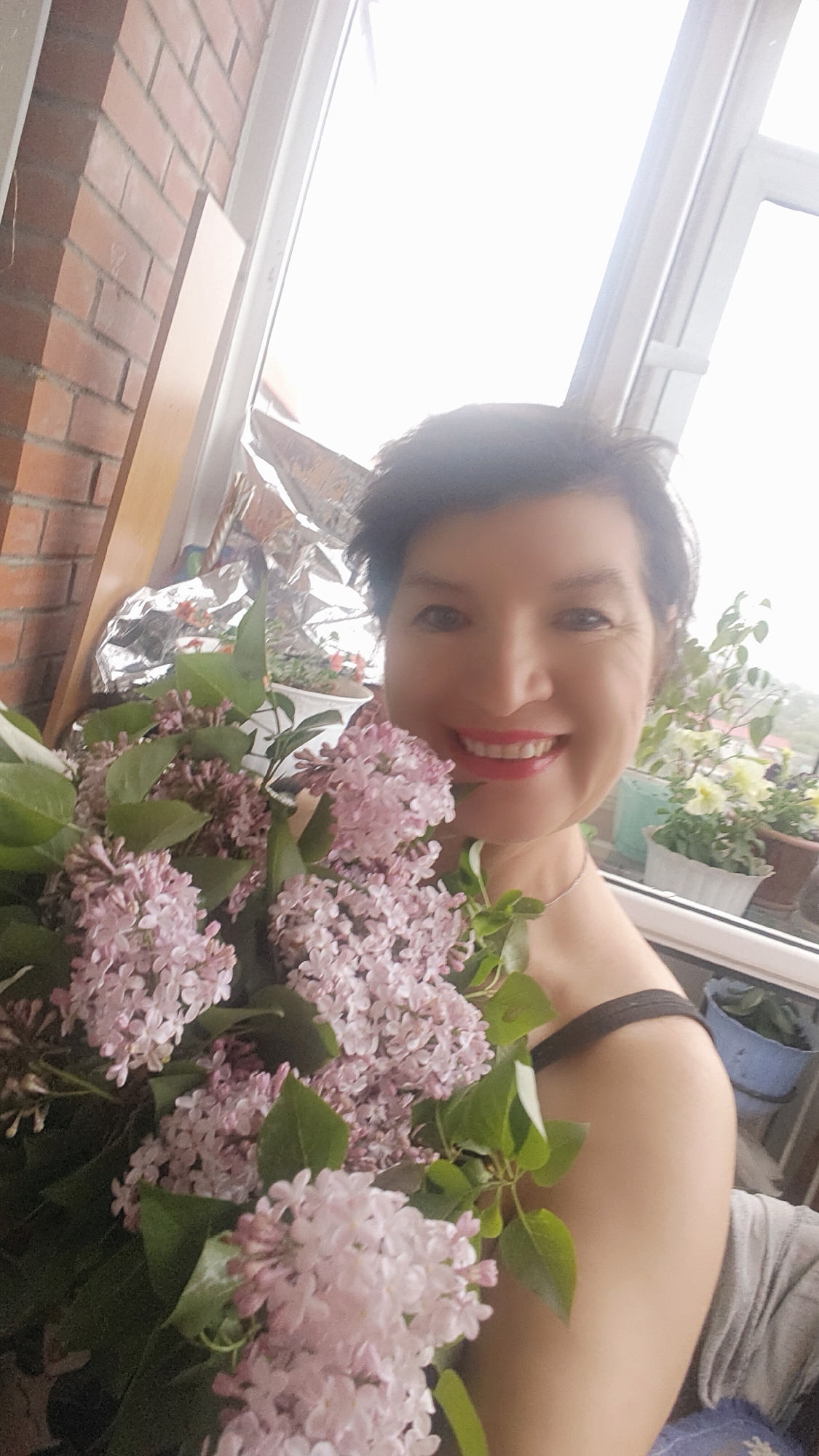 A woman smiles broadly, holding a large bouquet of light purple lilacs. She stands near a window, with a brick wall and various potted plants visible in the background. The location is likely a balcony or indoor space in Krasnoyarsk, Russia, based on the appearance of the building exterior and the type of plants commonly grown in the region. She is wearing a black top or dress.