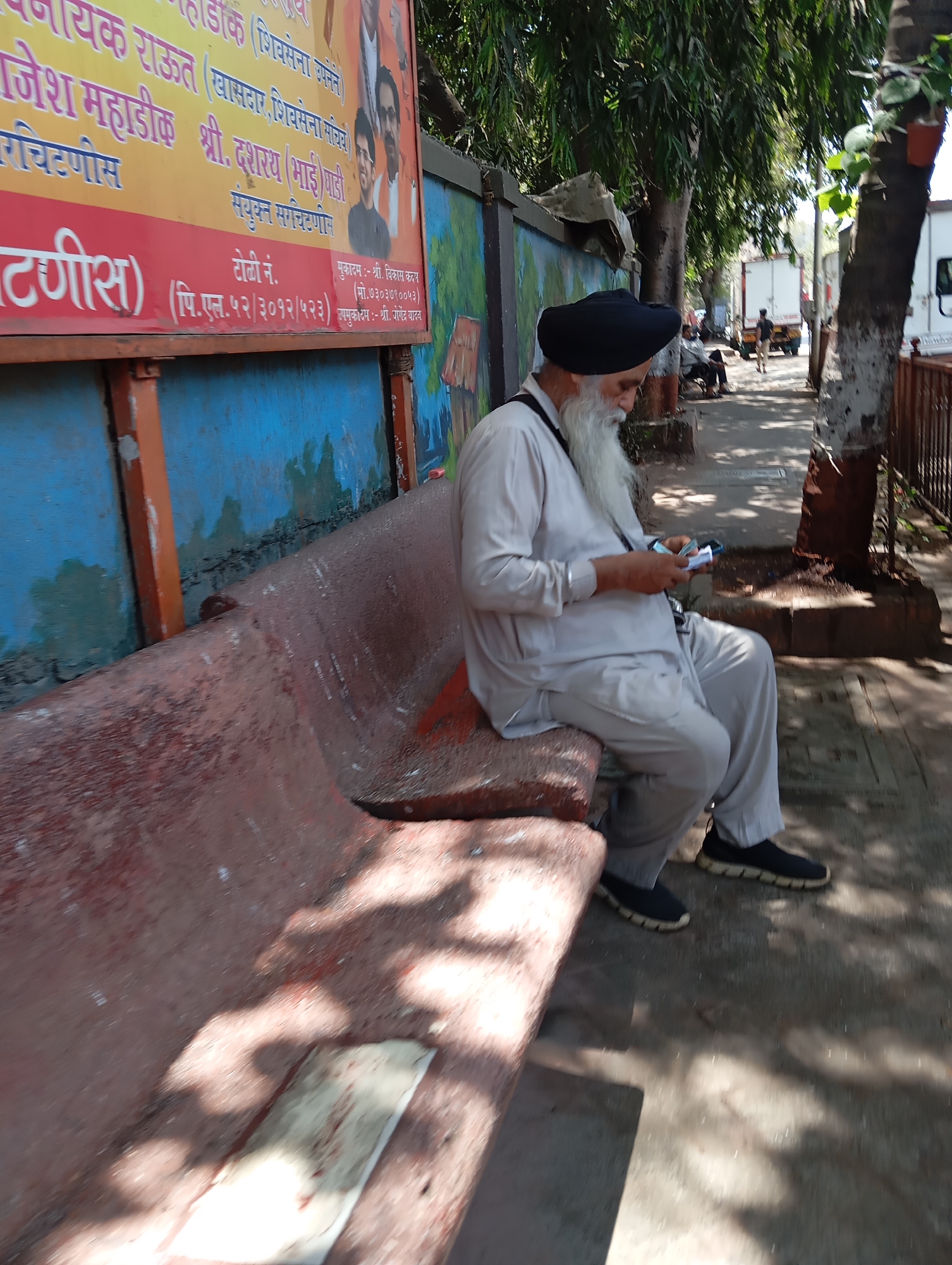 An older man with a long white beard, wearing a black turban and light-colored clothing, is sitting on a red bench while looking at something in his hands. Behind the bench is a mural or sign with text in what appears to be Marathi. The scene appears to be in Mumbai, India; the sign is likely a local political advertisement. The setting is outdoors, near a sidewalk and a tree. There is a person walking in the background and vehicles are parked along the street.