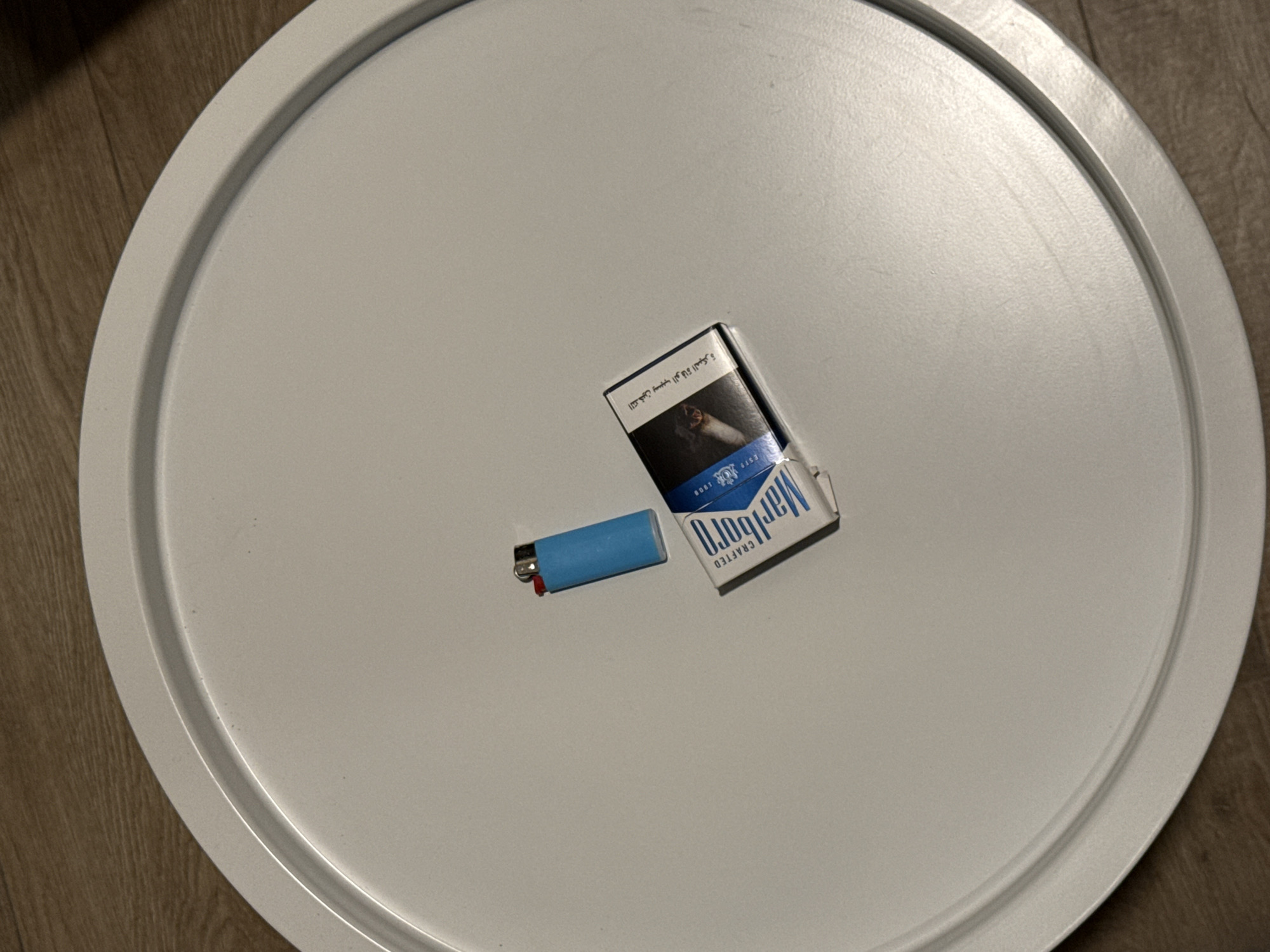 A blue disposable lighter and a pack of Marlboro cigarettes are placed on a white, circular tray, which is set on a wooden floor. The pack of cigarettes is partially open, revealing a few cigarettes inside. The pack has Arabic text printed on it, along with the Marlboro logo. The scene appears to be indoors, with no indication of the time of day or weather. The overall impression is one of everyday objects laid out on a flat surface, possibly for use. The setting of Dubai, United Arab Emirates, is provided as context.