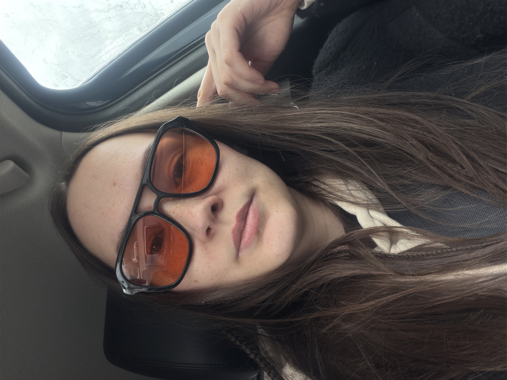 A woman is sitting in what appears to be the interior of a vehicle, possibly a car. She has long, brown hair and is wearing orange-tinted sunglasses. She is looking towards the camera, and her hand is positioned near her head. The vehicle's interior is visible, including the roof and seat. The location is unknown and there is no clear indication of Chaikovsky, Russia.