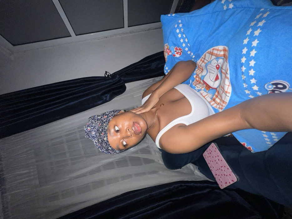 A young woman lies on a bed with a blue pillow featuring a cartoon character. She wears a white tank top and a patterned headscarf. She is tilted towards the camera. A mobile phone lies on her lap. The bed has a mosquito net draped around it and dark curtains are visible. The ceiling has a square pattern. The scene is dimly lit.