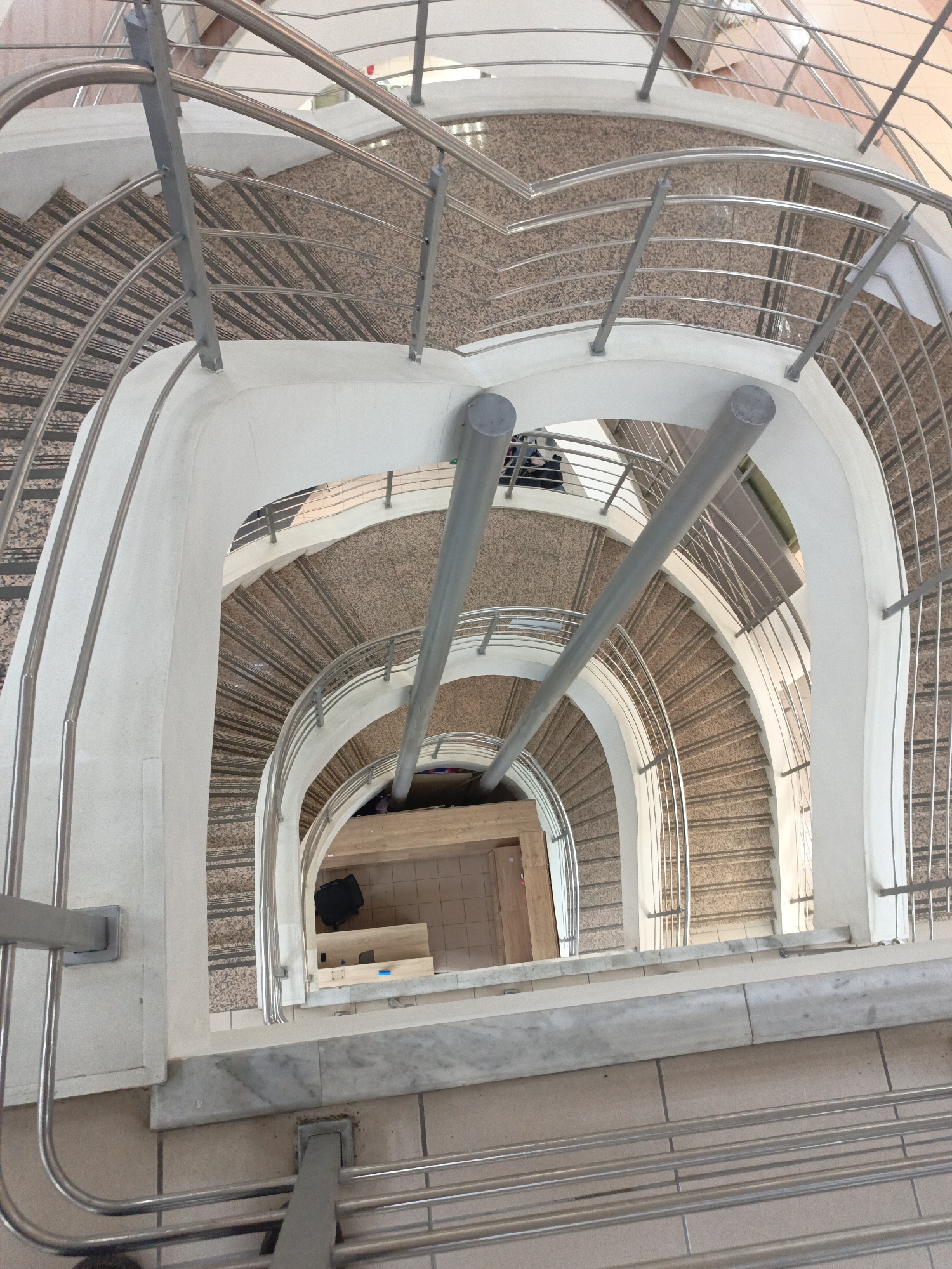 A high-angle, close-up shot depicts a spiral staircase within an architectural structure. The staircase's design incorporates a combination of white, curved walls and metal railings. The steps are covered with a speckled, beige and brown material. The bottom of the staircase reveals a section of a room with light brown tiled flooring and furniture, including what appears to be a box. Individuals, likely on different levels of the building, are partially visible through the staircase openings. The location is unspecified.