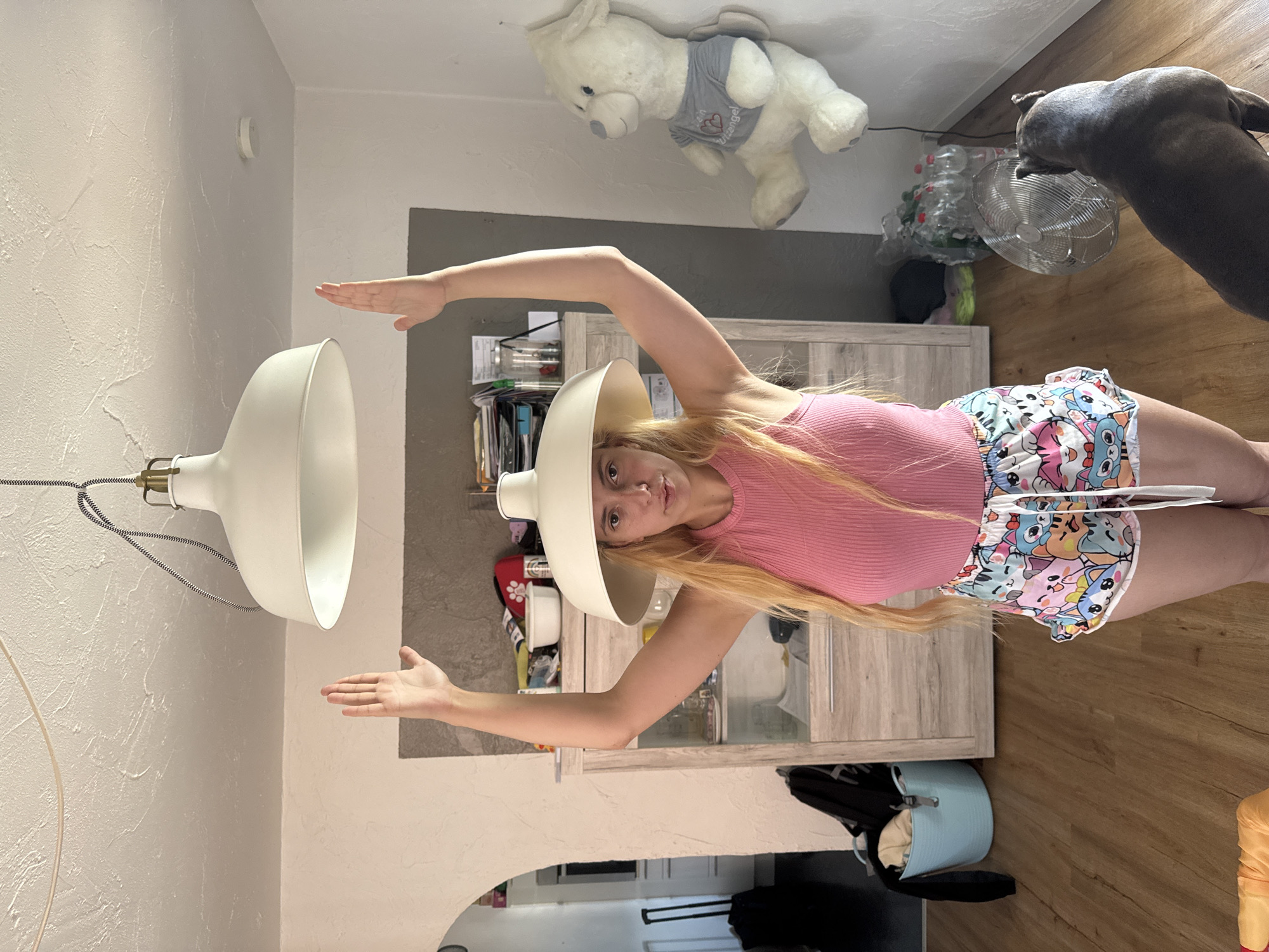 A young woman is standing in what appears to be a living room, wearing a pink tank top and shorts with a cartoon cat print. She has a lampshade on her head, and her arms are raised, holding a second lampshade above her head. A dark-colored dog is looking at her from the right side of the frame. In the background, there is a cabinet filled with various items, a large white teddy bear is propped up against the wall, and a fan is on the floor. The room has wooden floors and white walls with textured plaster. The lighting suggests it is daytime.