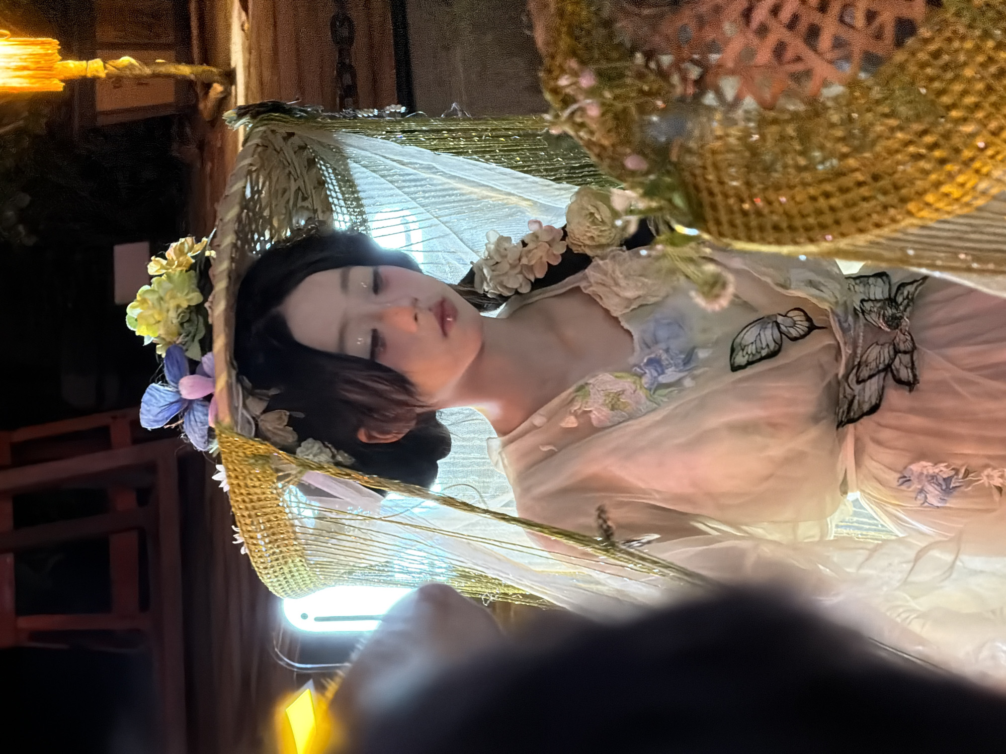 The image shows a close-up of a woman with dark hair and pale skin, lying down in what appears to be a theatrical setting. She is adorned with flowers in her hair and on her light-colored, sheer dress, which features embroidered butterflies. The woman has her eyes closed and her head tilted back, with a delicate, serene expression. The overall aesthetic is ethereal and dreamlike, possibly part of a performance or art installation. The background is dark and indistinct, with some wooden elements visible, suggesting an indoor or dimly lit outdoor environment. A bright, artificial light source is visible in the lower portion of the image. The composition is dominated by the woman's face and upper body, draped in flowing fabric and surrounded by decorative elements. The image was taken in Yuzhong District, China.