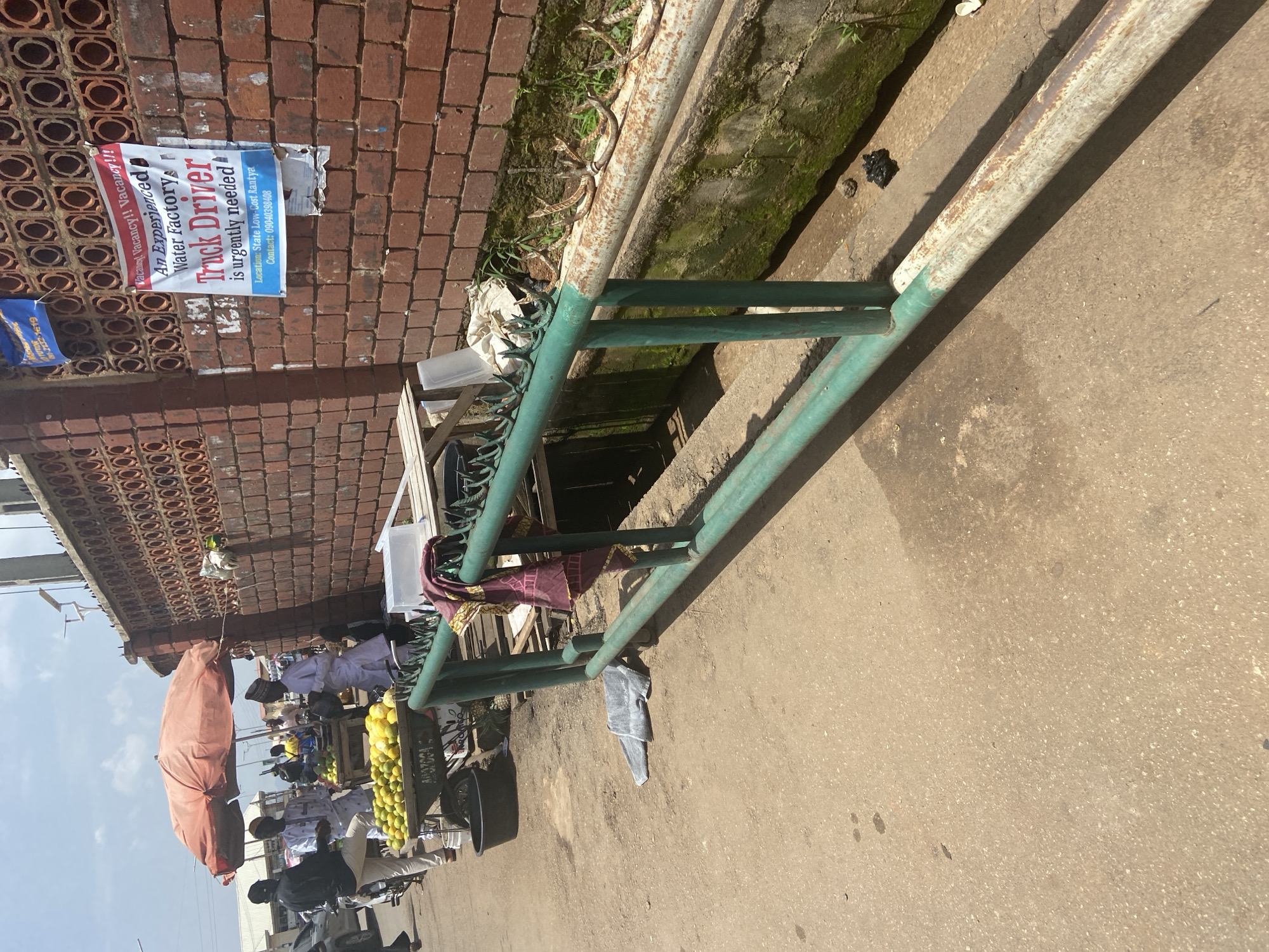 This is a medium shot of a street market scene in Jos, Nigeria, under a bright, sunny sky. In the foreground, a green metal railing with sections of rust and peeling white paint runs horizontally across the frame, separating the paved street from a drainage ditch. The ditch is partially overgrown with moss and weeds, and a rusty pipe runs along its edge.
To the left of the railing, a market stall is set up with a large, orange-red umbrella providing shade. Beneath the umbrella, a cart is filled with a large quantity of oranges and some other yellow fruits, possibly lemons or limes. Several people are gathered around the stall. One person, wearing white traditional attire and a cap, is standing near the cart, possibly a vendor. Another person in a black shirt and light-colored pants is standing next to a motorcycle, looking towards the stall. A third person in similar attire to the vendor is visible further back.
On the brick wall behind the market stall, several posters are affixed. One prominent poster, in white with red and blue lettering, advertises a "Vacancy" for an "Experienced Water Factory Truck Driver." It lists the location as "State Low-Cost Rantya" and provides a contact phone number. Another smaller blue poster with white lettering is visible to the left of this main poster.
The ground in front of the railing is dusty and paved, with some scattered debris, including a grey folded garment and some black plastic. The overall impression is a bustling, everyday scene in a commercial area of Jos.