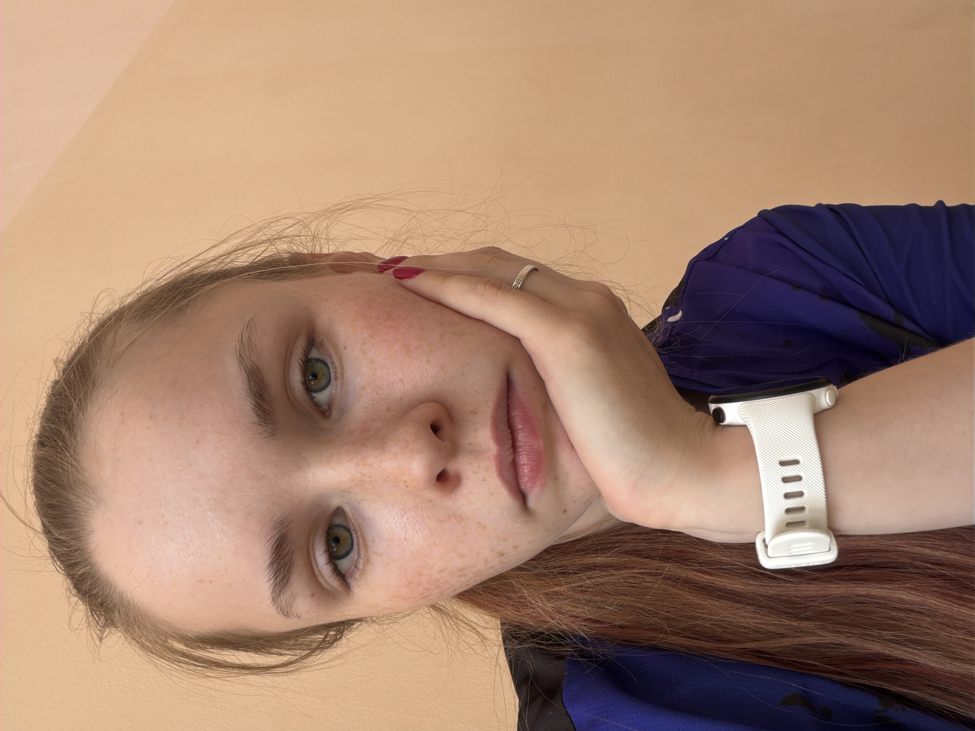 A close-up, portrait-style image features a young woman in an indoor setting, with a plain, light peach-colored wall as the background. The photograph is rotated 90 degrees clockwise, giving the impression that she is lying down and looking upwards towards the camera, or has her head tilted at a significant angle.
The woman has fair skin, distinctly adorned with numerous freckles across her nose and cheeks. Her eyes are a captivating green or hazel, maintaining a direct gaze towards the viewer. Her lips are naturally full and have a soft pink hue. Her hair, which appears to be light brown or auburn, falls around her face and extends towards the bottom right of the frame.
She is wearing a dark purple garment, possibly a t-shirt or sportswear. Her left hand is gently placed against her cheek, with her fingernails painted a deep red. A simple silver-colored ring with a small, subtle design is visible on her left ring finger. On her left wrist, she wears a white smartwatch featuring a ribbed strap. Her overall expression is neutral and calm. There is no visible text in the image.
