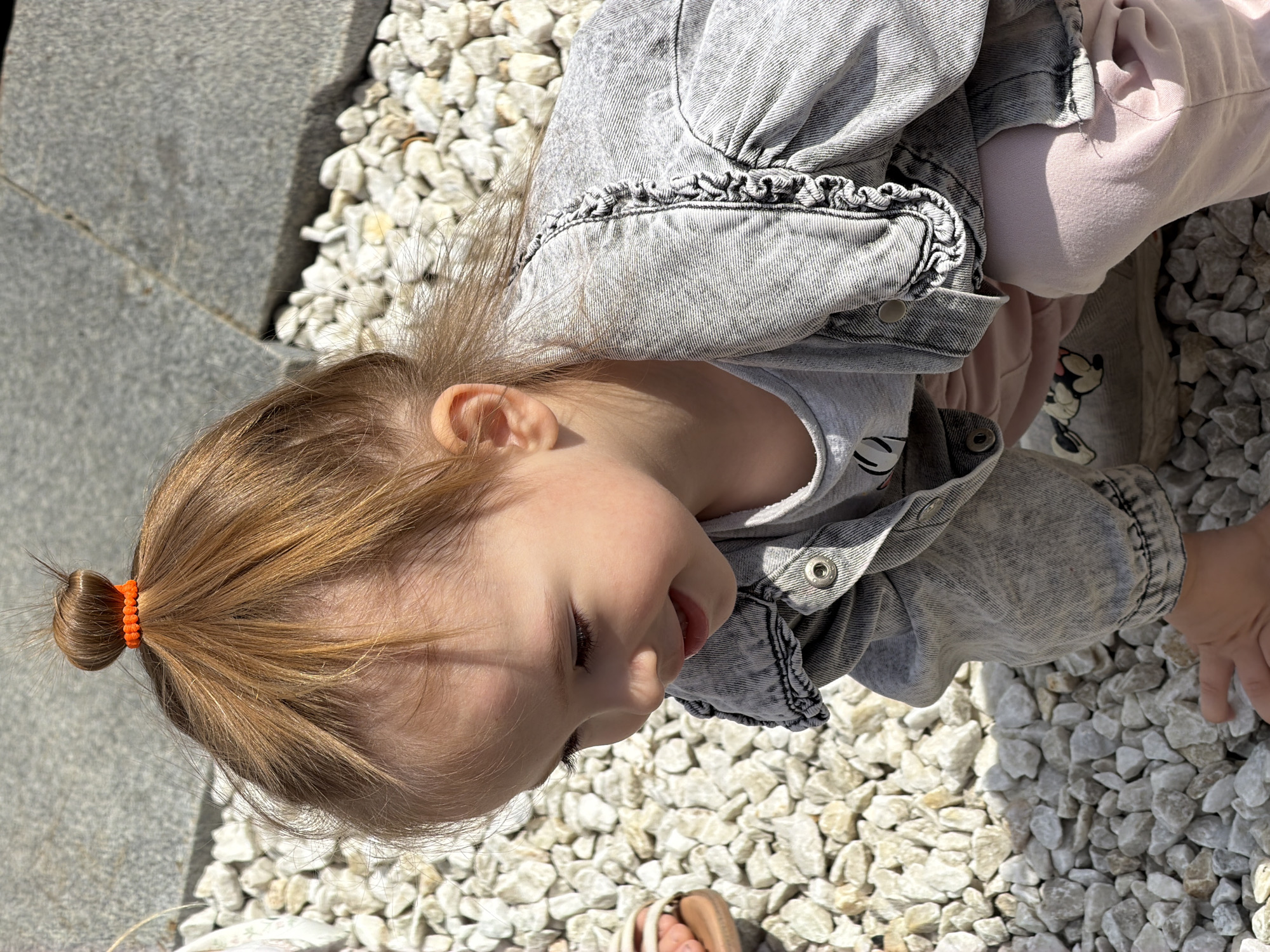A young child with auburn hair pulled into a top knot is crouching on a surface covered with white and grey gravel. The child is wearing a light grey denim jacket with ruffles along the collar and cuffs, and a light grey t-shirt underneath. The child's hands are visible near the gravel, suggesting interaction with the ground. In the background, large grey stones are visible. The lighting indicates it is daytime, with shadows cast on the gravel. The location is specified as Ufa, Russia.