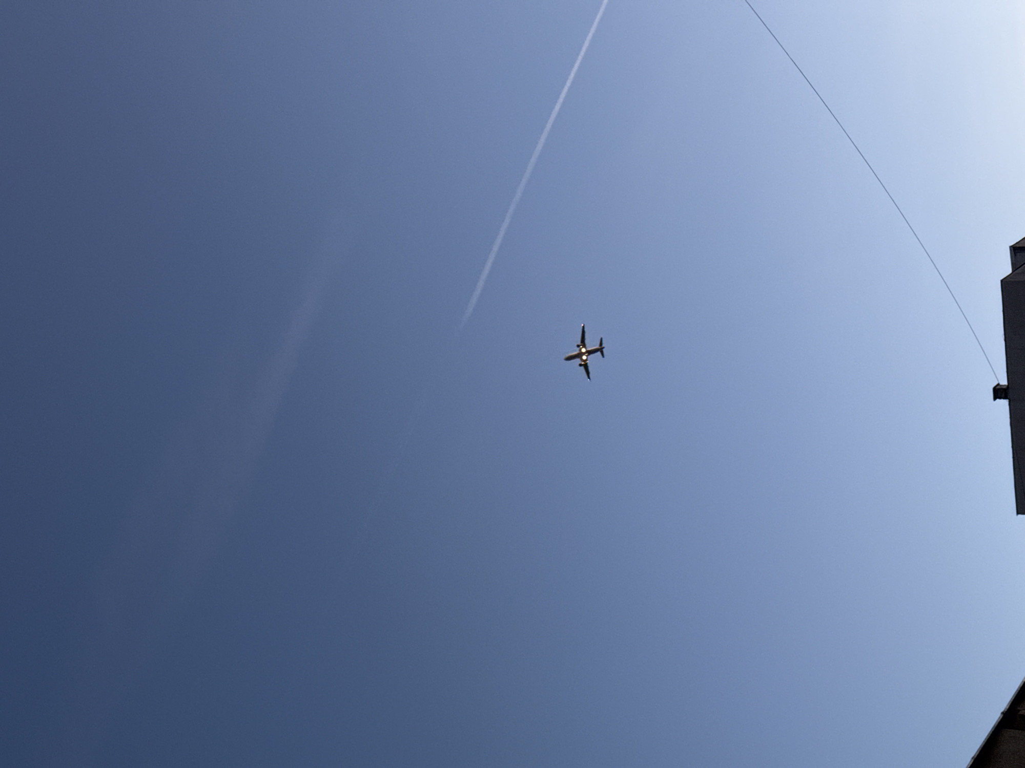 In the image, a passenger airplane is depicted flying across a clear blue sky, leaving a white contrail behind it. The plane is silhouetted against the bright sky, with its lights visible. In the foreground, the rooftops of buildings and a power line are visible, suggesting an urban setting. The overall scene is serene, capturing a moment of everyday life in Varna, Bulgaria, with the airplane serving as a symbol of travel and connection.