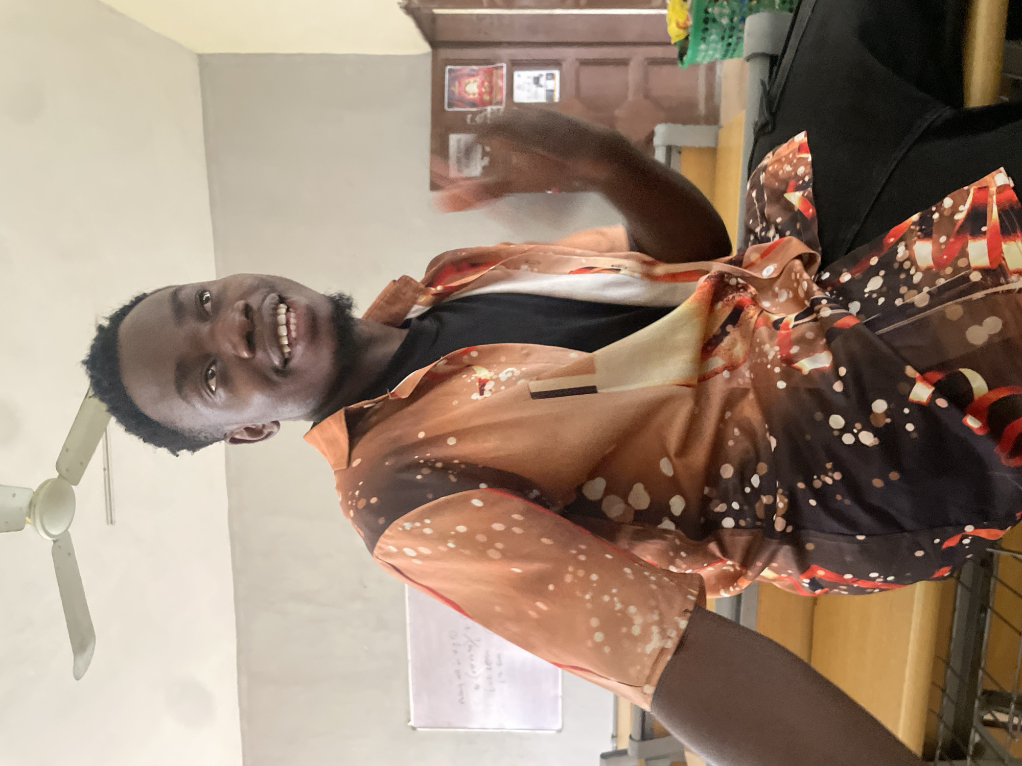 A young Black male is captured from the chest up, seated indoors. He is wearing a short-sleeved, collared orange shirt with a speckled pattern in white and brown, over a black t-shirt. His head is turned to the left, and he is smiling broadly, showing his teeth. His right hand is raised and partially blurred, suggesting movement. The background shows a classroom or lecture hall environment with a whiteboard on the left, chairs and desks, and a ceiling fan above. The lighting is diffused, suggesting an indoor setting possibly during the day. The overall impression is one of a casual and engaging moment. The location is identified as Ihiagwa, Nigeria.