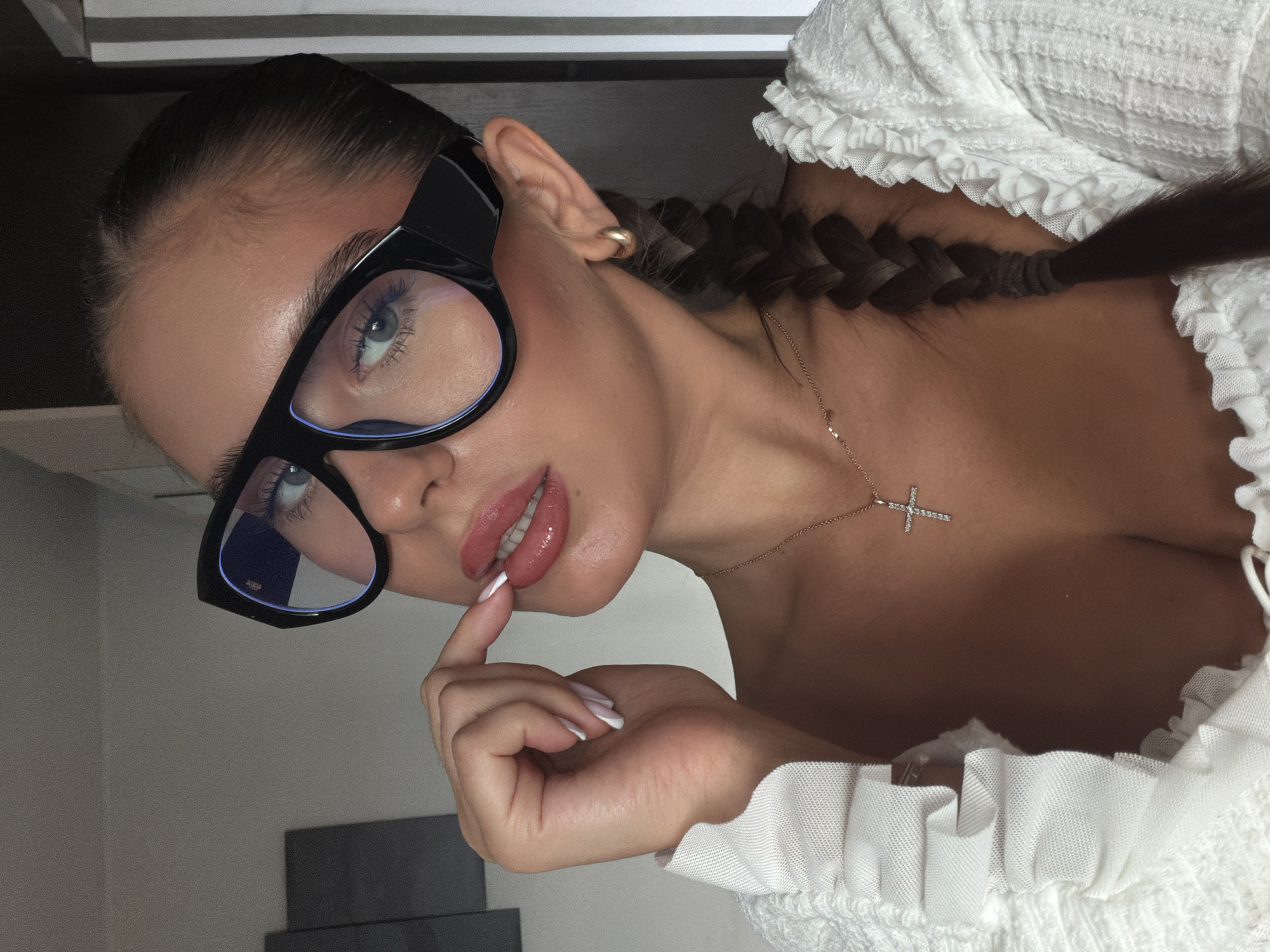 A young woman with fair skin, styled dark hair in a braid, and wearing black-rimmed glasses is the primary subject of the image. She is positioned facing the camera, with her gaze directed slightly upwards. Her right hand is raised, with a finger touching her lip. She is wearing a white top with ruffled detailing. A gold necklace with a cross pendant is visible. The background is a simple off-white wall, with a dark, rectangular object visible in the lower left corner. The location is not discernible from the visual information provided.