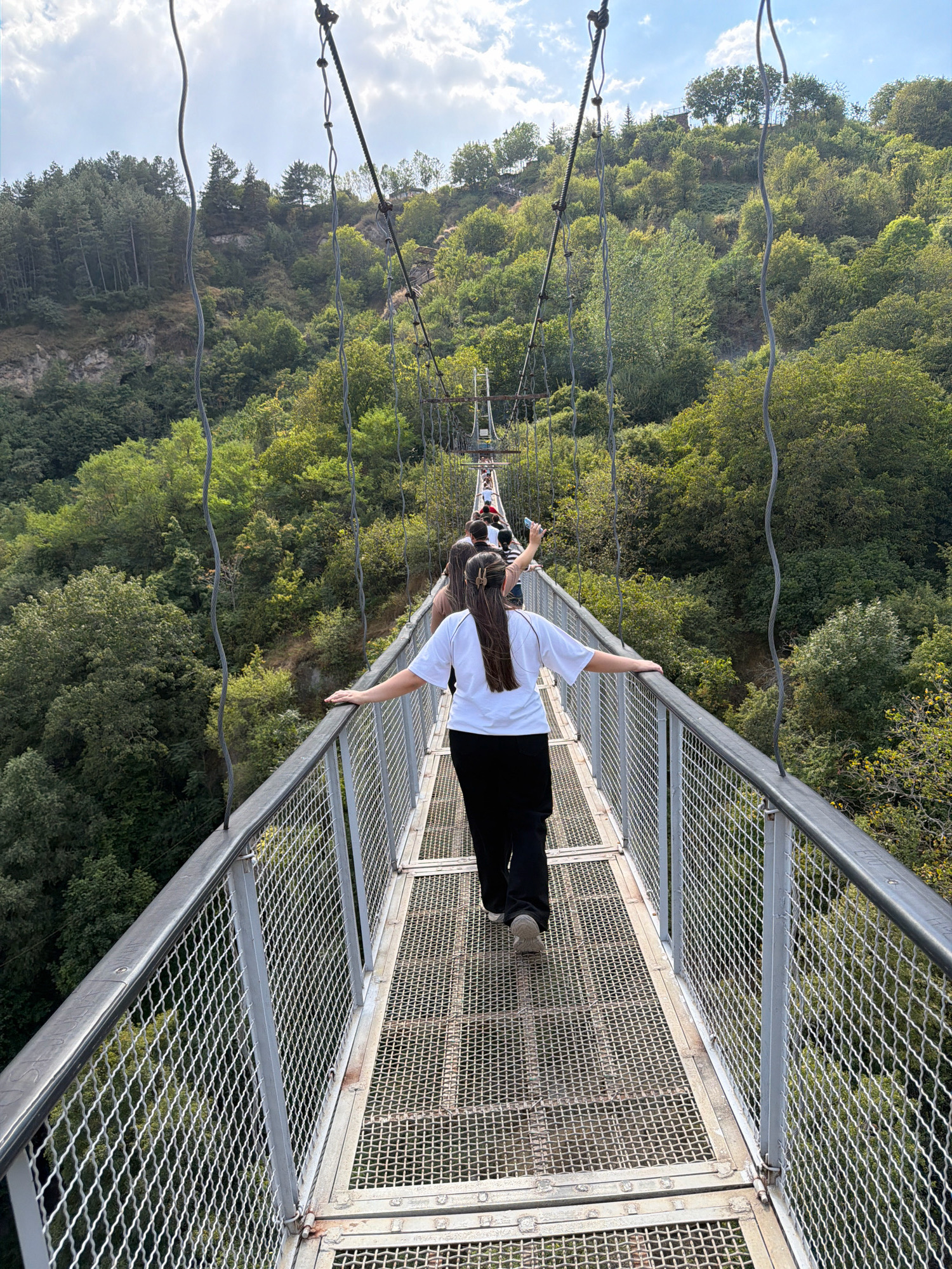 The image shows a long, narrow suspension bridge spanning a green, wooded gorge. The bridge has metal railings and a metal mesh walkway. Several people are walking on the bridge, with the closest person in the foreground facing away from the camera, wearing a white t-shirt and black pants. The bridge is suspended by thick cables, and the surrounding environment is lush with trees and vegetation. In the distance, the bridge continues, with more people walking on it, and appears to lead to a hillside with trees. The sky above is partly cloudy. This is the Khndzoresk Suspension Bridge in Khndzoresk, Armenia.