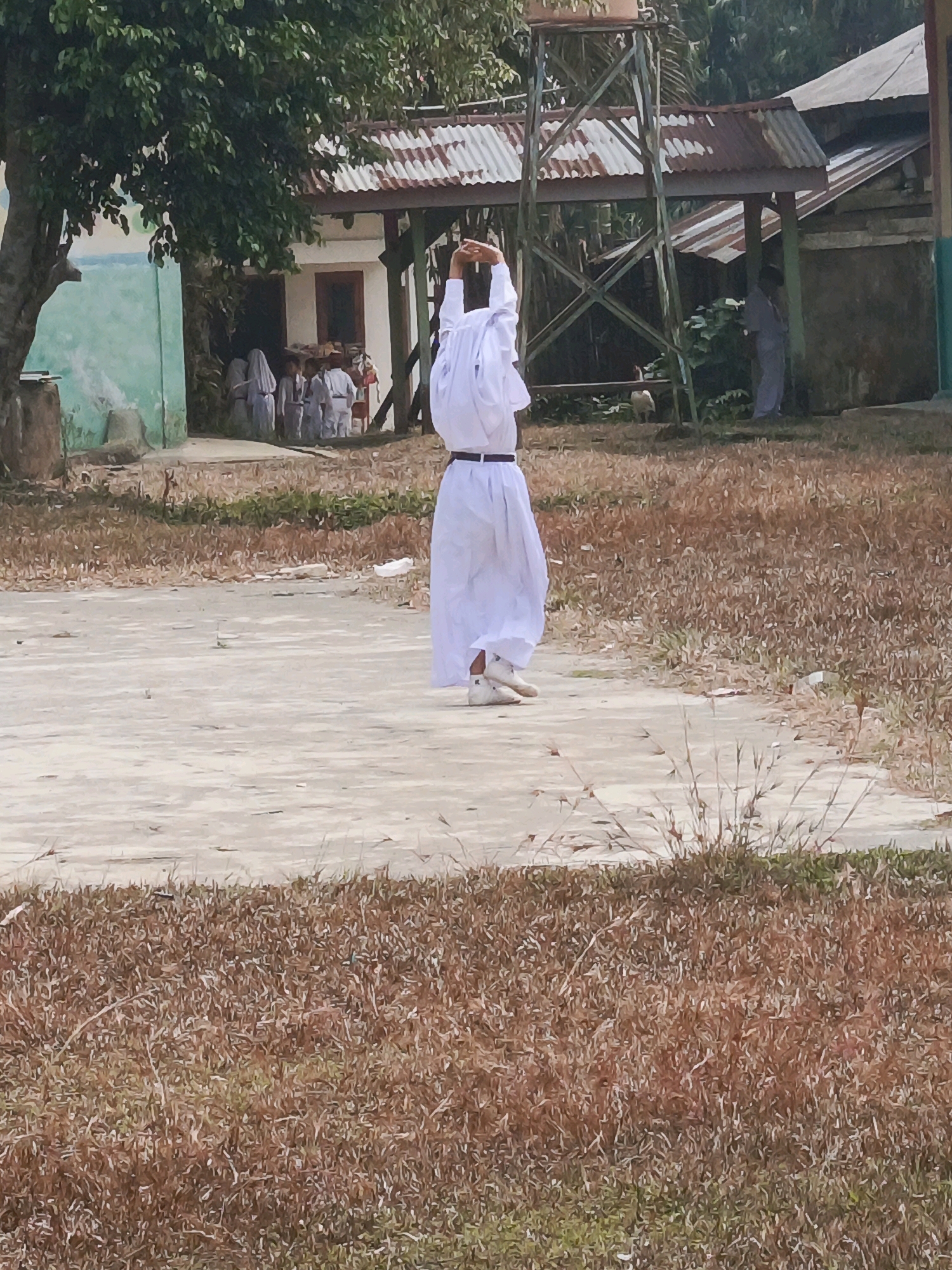In a dry, grassy outdoor area in Wonosari, Indonesia, a person in a white, full-length robe and white sneakers stands facing away from the camera with arms raised overhead. To the left of this central figure, near a green-painted wall and under a large tree, a group of individuals in similar white attire stand in a semi-circle, appearing to be observing or participating in an activity. Behind them, a building with a corrugated metal roof and a water tower are visible. To the right of the central figure, a person in a grey outfit stands near another covered structure. The ground is a mixture of dry grass and a concrete-like surface.