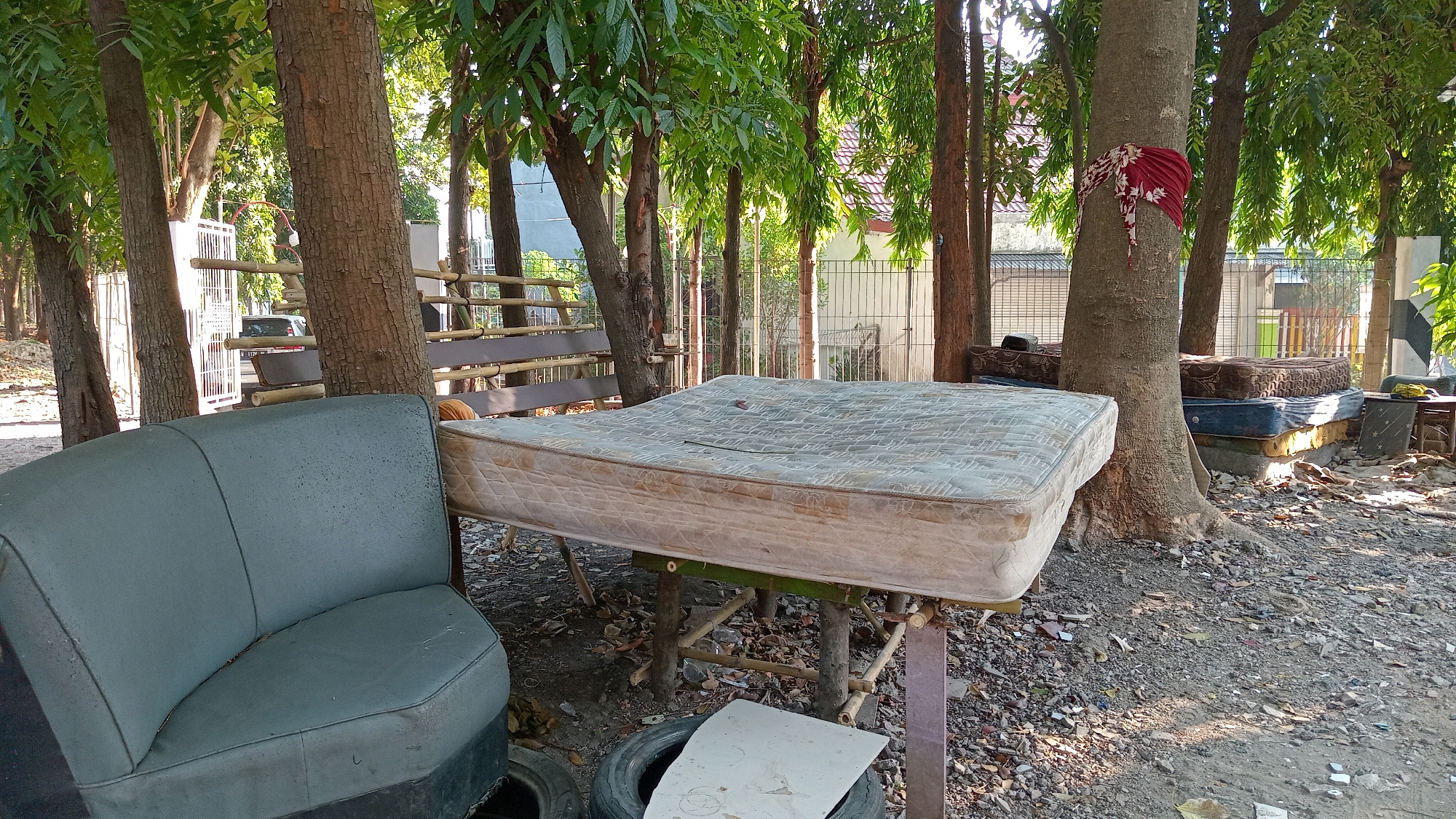 The image depicts an outdoor scene in Randuagung, Indonesia, characterized by a collection of discarded mattresses and furniture arranged amidst trees and gravel. In the foreground left, a faded teal-colored, two-seater bench or sofa, likely from a vehicle, is positioned on top of a tire. Dominating the center is a large, soiled mattress resting on a makeshift table constructed from bamboo and planks. To the right of this, several more mattresses are stacked. A large tree with a red and white cloth tied around its trunk stands prominently in the mid-ground, with more stacked mattresses behind it. The environment is littered with debris, including broken pieces of tile and leaves. In the background, a white metal fence and buildings are visible, suggesting the location is near residential or commercial areas. The overall impression is one of an informal outdoor storage or disposal area for used bedding and furniture.