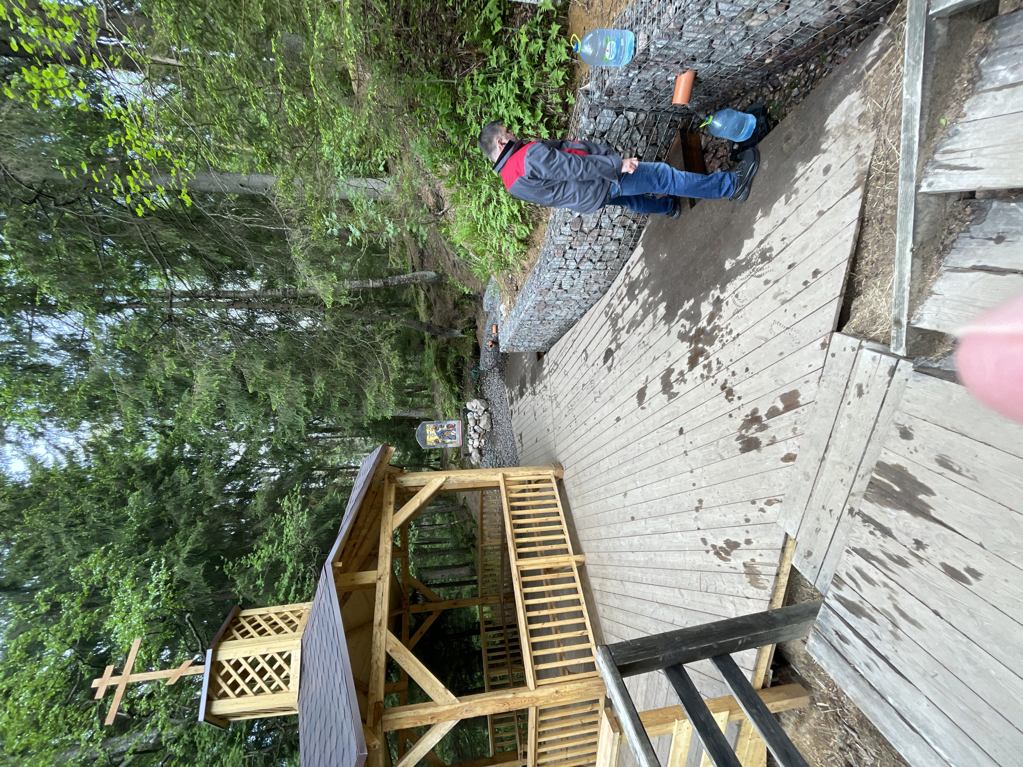 A man stands on a wooden platform in a wooded area of Unknown, Russia. He is wearing a gray jacket with red accents and blue jeans, and he is filling a large blue water bottle from a pipe extending from a stone wall. Next to the platform, there is a wooden structure with a cross on the top, suggesting it is a religious site or chapel. The wooden platform appears damp with spots of water. Trees dominate the background.