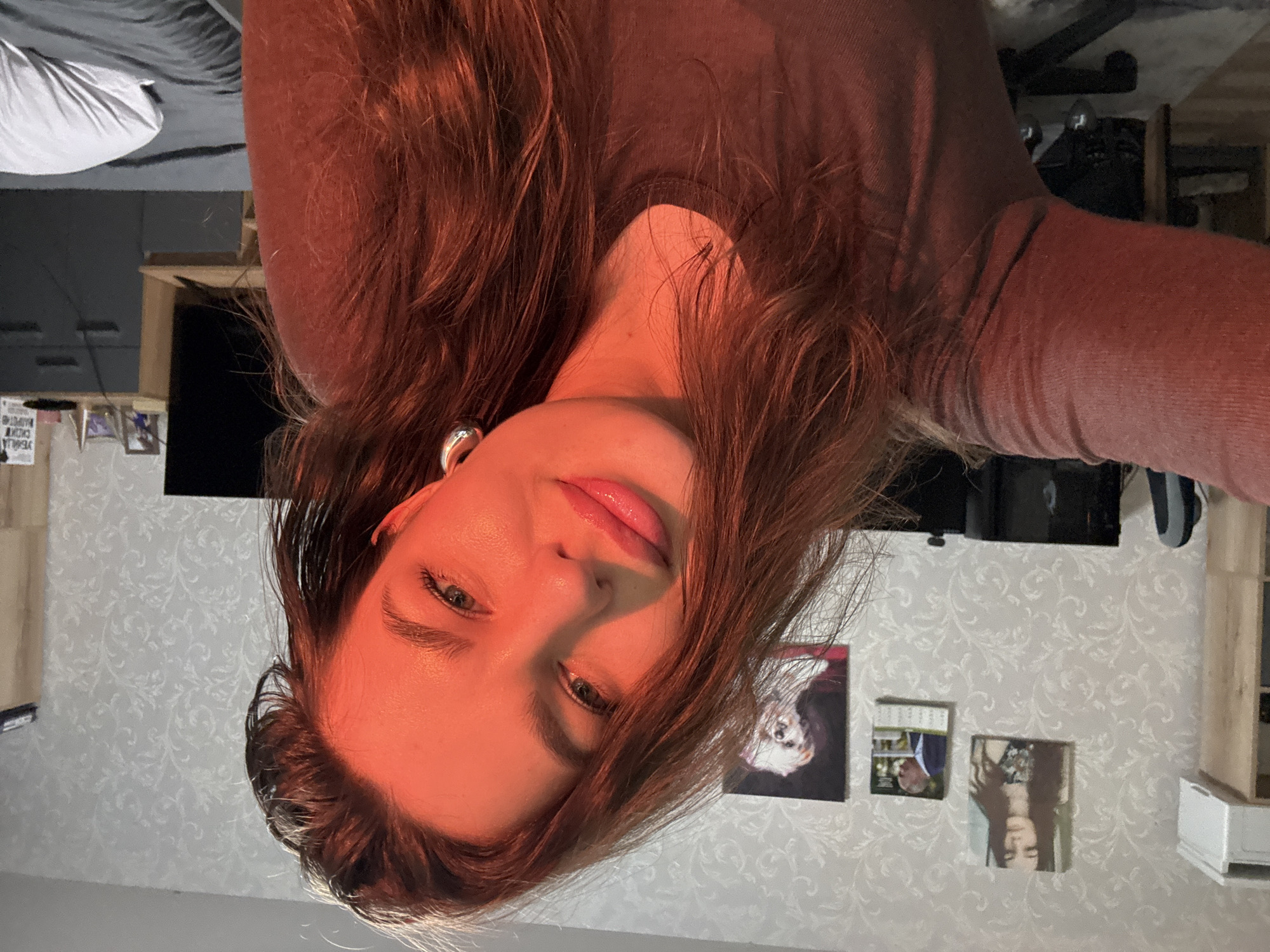 In the image, a young woman with long, wavy brown hair is captured in a selfie. She is wearing a brown long-sleeved shirt and large silver hoop earrings. The lighting in the image is warm, with a reddish hue, suggesting either sunset or artificial lighting. The woman's expression is relaxed and slightly playful, with a subtle smile and her eyes looking towards the camera.
The background appears to be an indoor setting, possibly a bedroom or living area. There are framed pictures on the wall, including one of a woman, a man, and a dog. Behind her, a television screen is visible, along with furniture such as a desk and a cabinet. The wallpaper has a subtle floral pattern.
While the image is set in Russia, the specific location within the city of Chaikovsky is not discernible from the visual information provided.