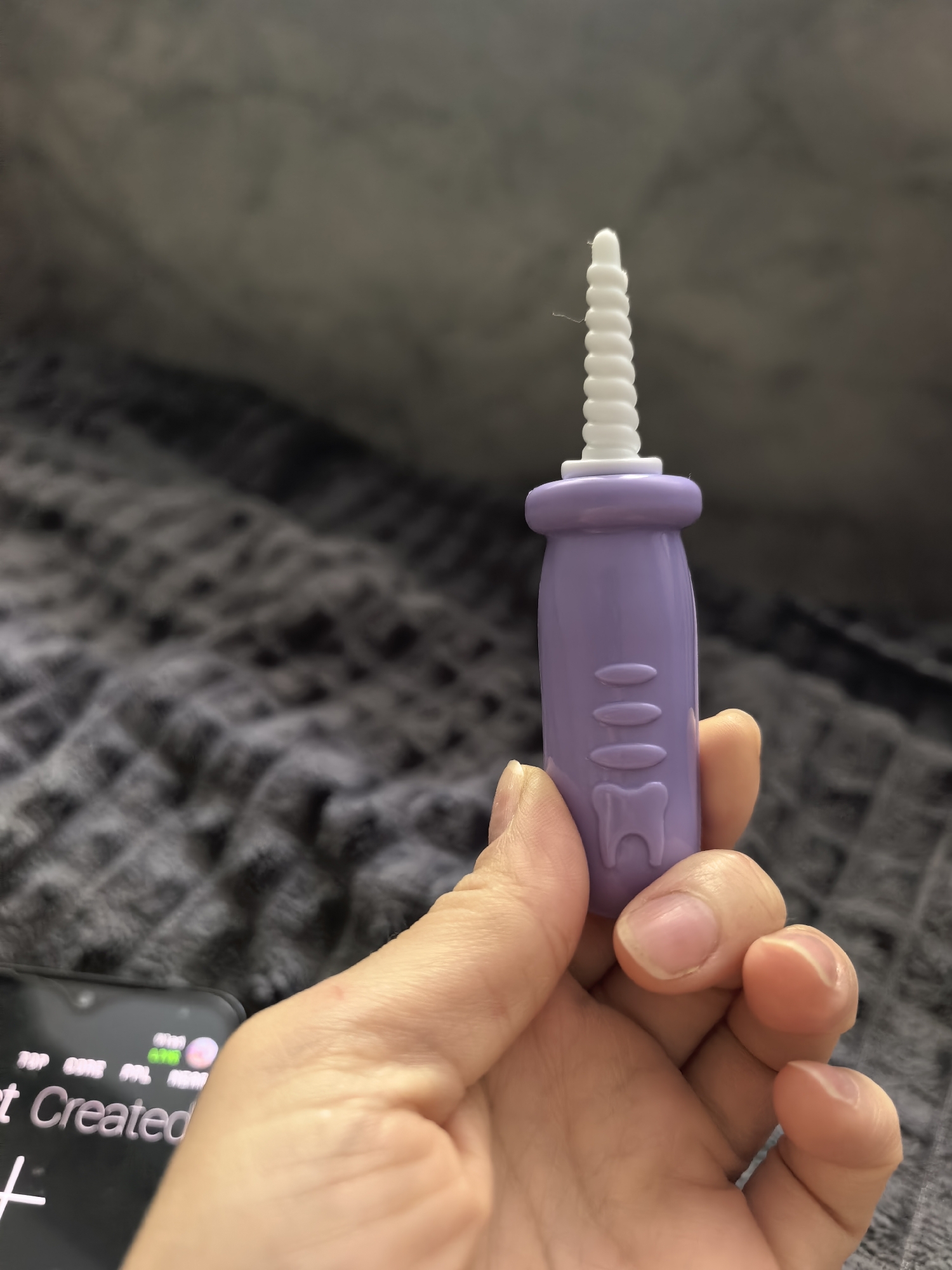A fair-skinned hand is holding a purple plastic dental tool with a white spiral-shaped brush head. The tool has three horizontal oval indentations and a tooth-shaped design near the bottom. A black smartphone is partially visible on the left side of the frame. The background consists of a gray textured blanket. It is not possible to determine the location as Sterlitamak, Russia from this image.