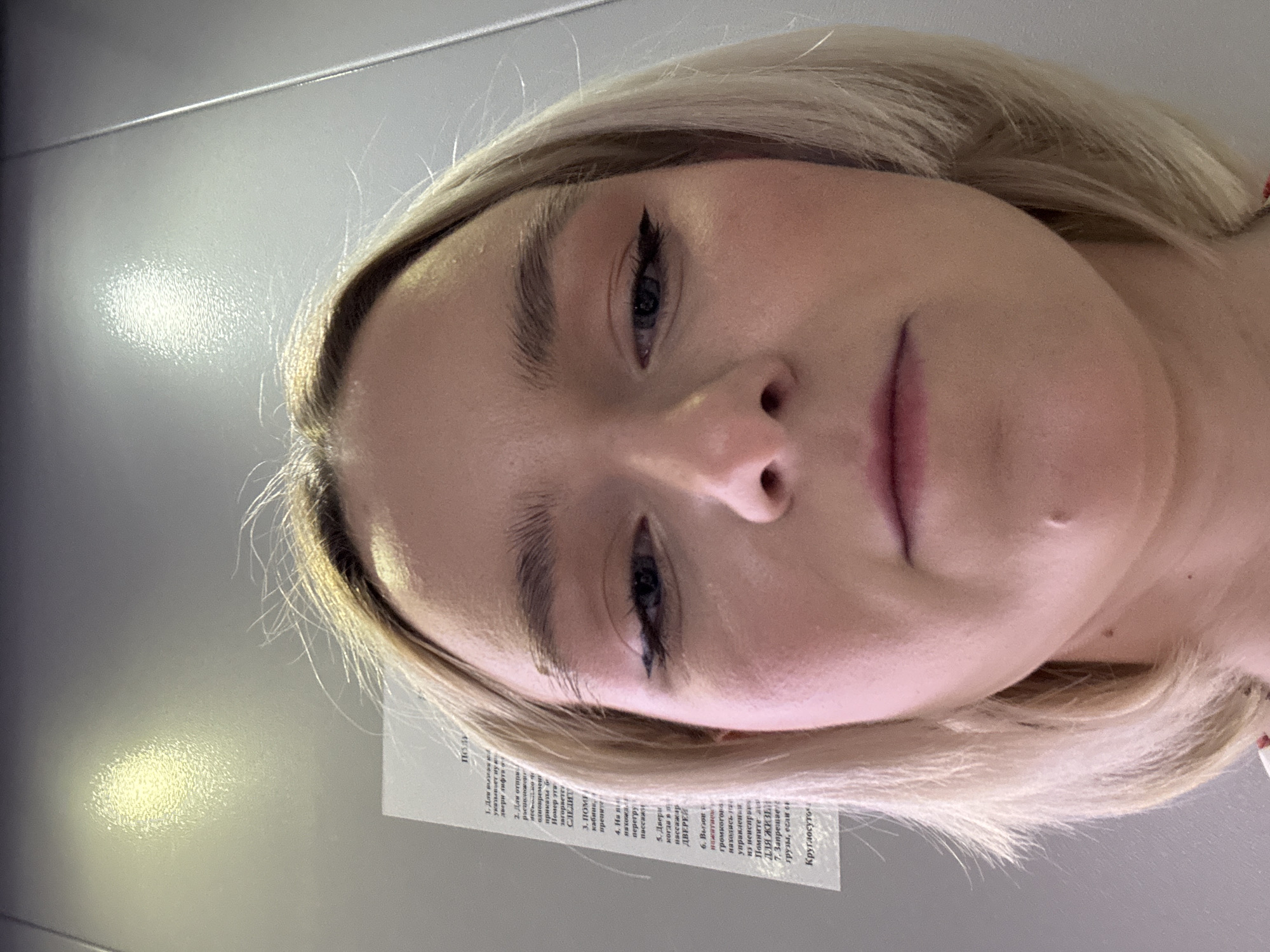 The image is a close-up selfie of a blonde woman with black eyeliner. A piece of paper is visible in the lower left corner, with text written in Russian. The paper includes a title "Пол" (floor), followed by a numbered list of instructions, possibly related to elevator usage. The last word in the Russian text on the paper, visible in the lower right corner, is "Круглосуто" (round the clock). The location cannot be determined without more information.