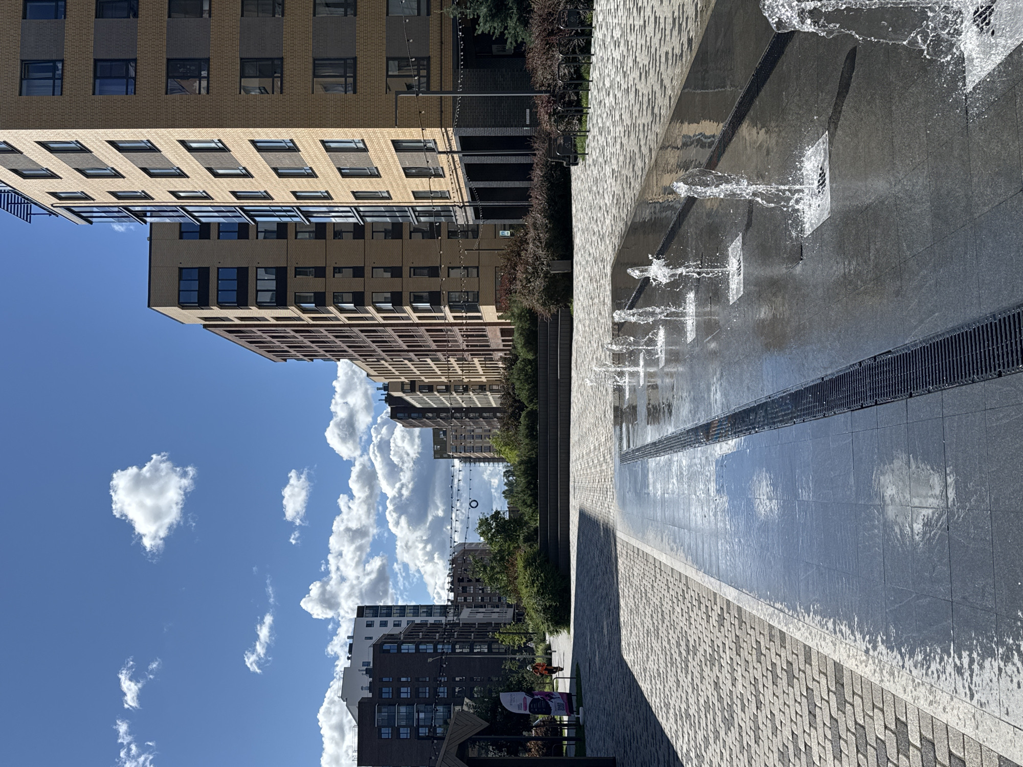 A sunlit, modern urban courtyard in Novoselye, Russia, is captured on a clear day with scattered white clouds. The foreground features a rectangular water feature with multiple jets of water spraying upwards from square openings set into a dark, reflective tiled surface. The water creates splashes and mist, with reflections of the sky and surrounding buildings visible on the wet tiles. A dark grate runs along the edge of the water feature. To the left of the water feature is a paved walkway constructed of light-colored, textured rectangular stones arranged in a herringbone pattern.
Beyond the walkway, a grassy area with manicured bushes and small trees lines the path. In the middle ground, several contemporary multi-story apartment buildings are visible. These buildings exhibit varied architectural styles, with some featuring yellow brick facades and numerous windows, while others have darker brick and glass exteriors. A person wearing an orange safety vest and a white hard hat is walking on the paved path towards the left side of the frame, carrying a piece of equipment. A large, colorful banner is also visible near the person. In the distance, more buildings and trees extend into the scene. The sky above is a vibrant blue, dotted with fluffy white clouds.