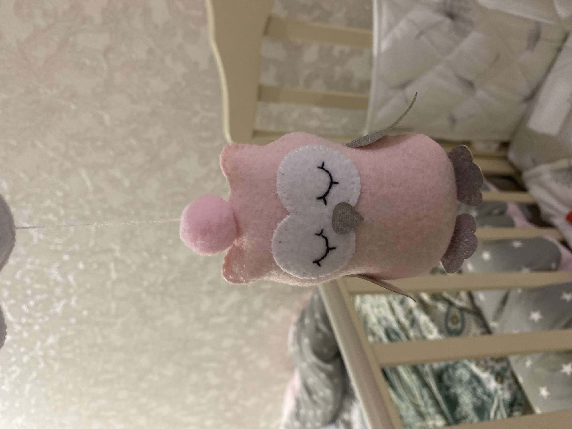 The image captures a pink felt owl toy suspended by a thin white string, slightly out of focus, within a nursery in Tyumen, Russia. The owl, constructed from felt, presents a pink body with white facial features depicting closed eyes with black embroidered eyelashes, a grey beak, and grey feet. Small grey felt wings protrude from its sides, and a pink pom-pom is affixed to the top of its head. In the left background, a portion of a light-colored crib is visible, containing patterned bedding in green and white paisley, alongside grey fabric with white stars. A grey textile item, possibly a blanket or pillow, rests over the left crib railing. The right background reveals another section of a light-colored crib, featuring a white padded crib bumper or mattress cover. The wall behind the owl is covered with a light-colored wallpaper displaying a subtle, textured, and diffused pattern. Illumination is consistent with interior artificial light.