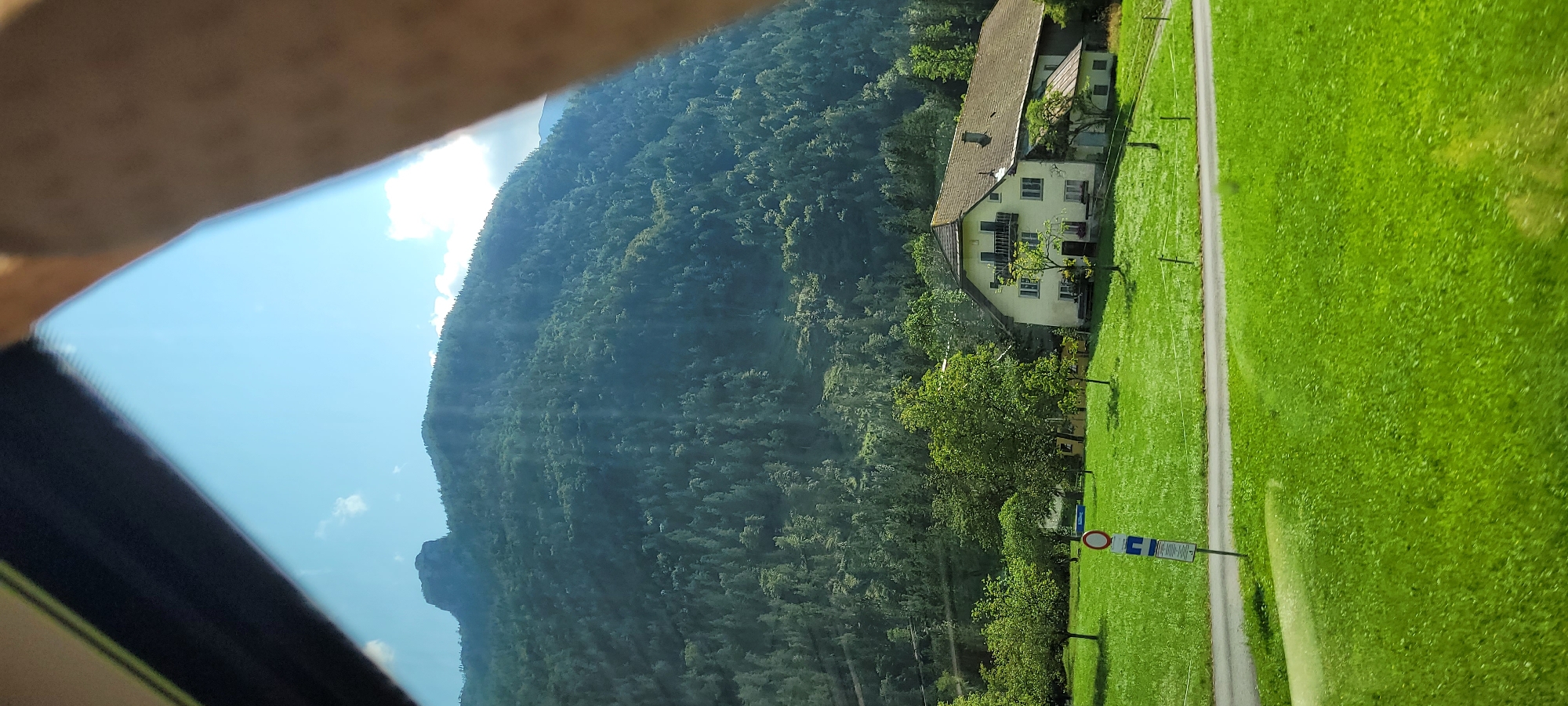 In a bright, sunlit outdoor scene, a forested mountain dominates the background under a clear blue sky with scattered white clouds. The mountain's slope is densely covered with evergreen trees, with a prominent, rocky peak visible at the summit. In the mid-ground, a traditional Austrian farmhouse with a light-colored facade, dark-tiled roof, and balconies stands on a lush green lawn. Several mature trees surround the house. A paved road curves through the lawn in front of the house, and a road sign with a circular red and white symbol and a rectangular blue sign below it is placed on the side of the road. The foreground consists of a vibrant green, well-maintained lawn, suggesting a rural or semi-rural setting. The image is partially obscured by a dark-colored frame in the upper left and a blurred human arm in the upper right, indicating the photo was taken from inside a vehicle or through a window. The location is identified as Georgenberg, Austria.