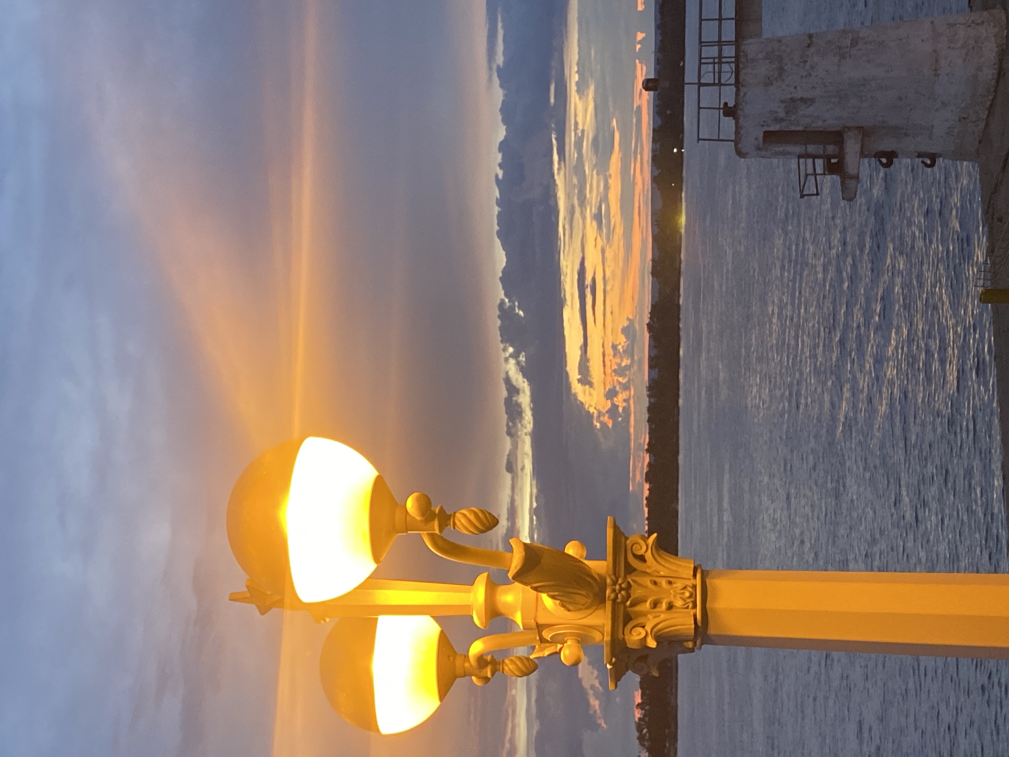 This is a photograph taken during sunset in Novokuybyshevsk, Russia. In the foreground, a tall, ornate, golden lamppost with two lit globes is positioned to the left of the frame. The lamppost has a fluted column and decorative scrollwork. Behind the lamppost, a wide body of water, likely a river, fills the middle ground. The water is calm and reflects the colors of the sky. In the background, a distant shoreline with trees is visible. The sky is a dramatic mix of colors, with oranges, yellows, purples, and grays, indicating the sun is setting. On the right side of the frame, a portion of a concrete structure, possibly a pier or a lighthouse foundation, is visible. There are no people or animals visible in the image. The overall scene is peaceful and atmospheric, capturing the transition from day to night.