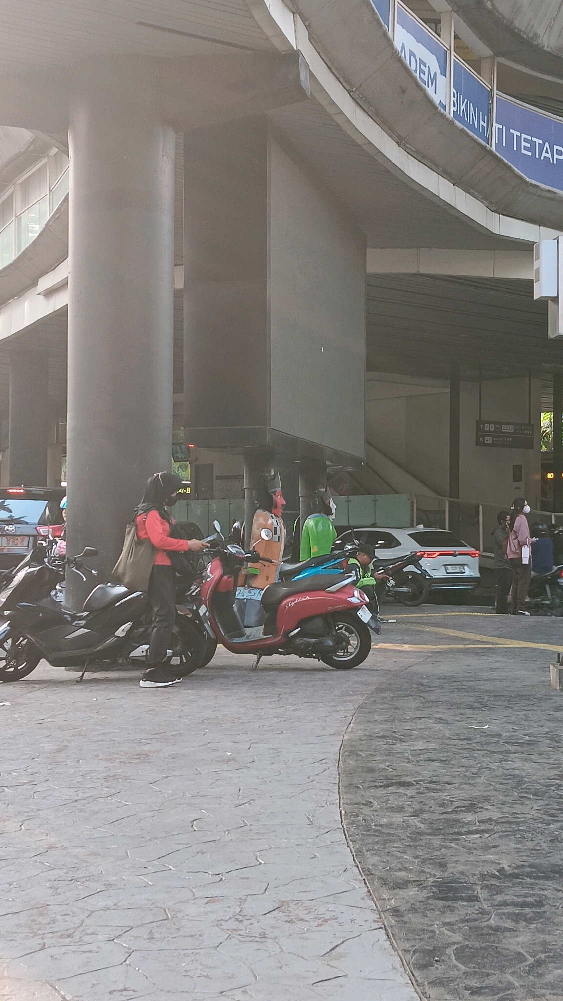 A street scene in South Jakarta, Indonesia, features a woman in a red shirt and black hijab standing next to a red scooter. She is looking at her phone. To her left is a black scooter. Behind the scooters are two large figures, one in orange and the other in green. Several other people and vehicles are visible in the background. The ground is paved with stone tiles. An elevated walkway or bridge is visible overhead with signage.