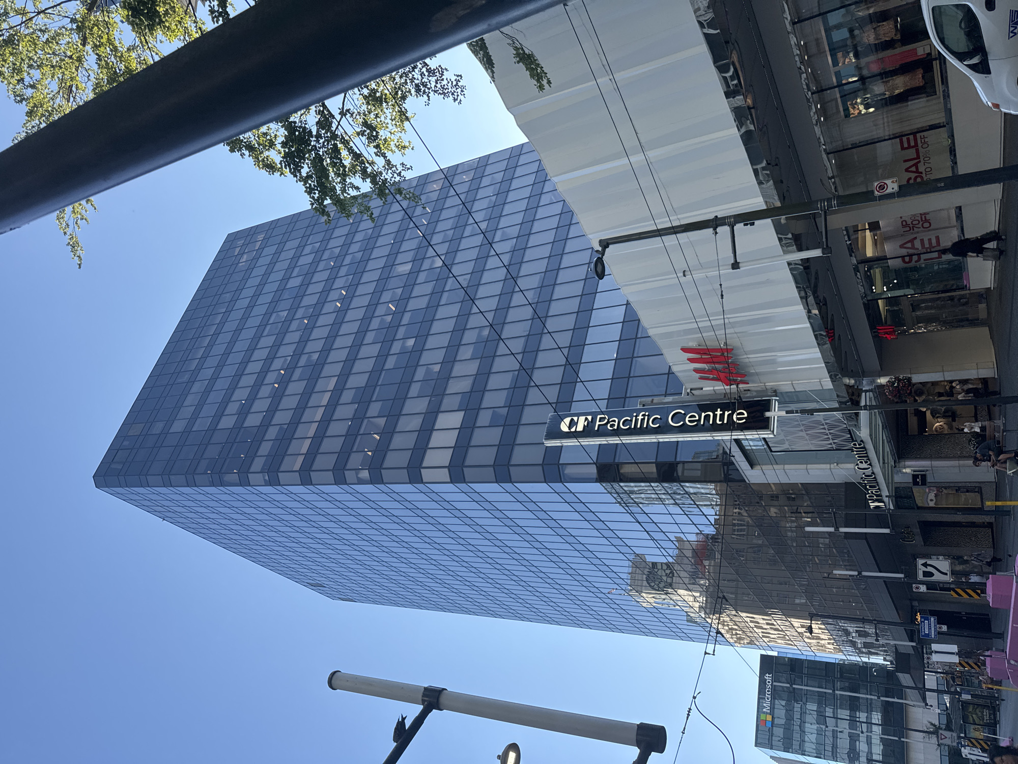 The image shows a street scene in Vancouver, Canada, dominated by a tall, modern glass skyscraper reflecting the clear blue sky. In the foreground, a street level storefront with "H&M" signage is visible, featuring large display windows with "SALE UP TO 70% OFF" text. To the left of the skyscraper, another building with a prominent "Microsoft" logo can be seen. The street is lined with utility poles and wires, and several people are walking on the sidewalk. The overall environment suggests a busy urban commercial district.