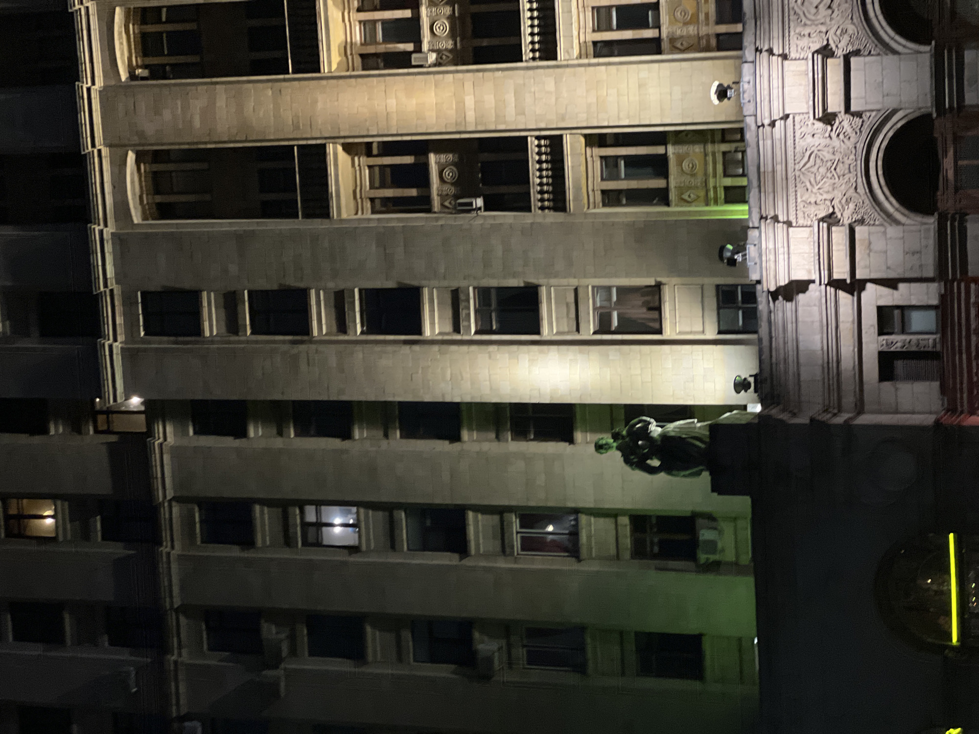 Captured at night, the image portrays a close-up view of a building, likely situated in the city of Moscow, Russia. The building's architecture is characterized by multiple floors with distinct rows of windows, some displaying interior lights. A sculptural element is visible on the building's facade. The illumination casts shadows and highlights the building's textural details. A street lamp's yellow reflection is visible at the bottom right.