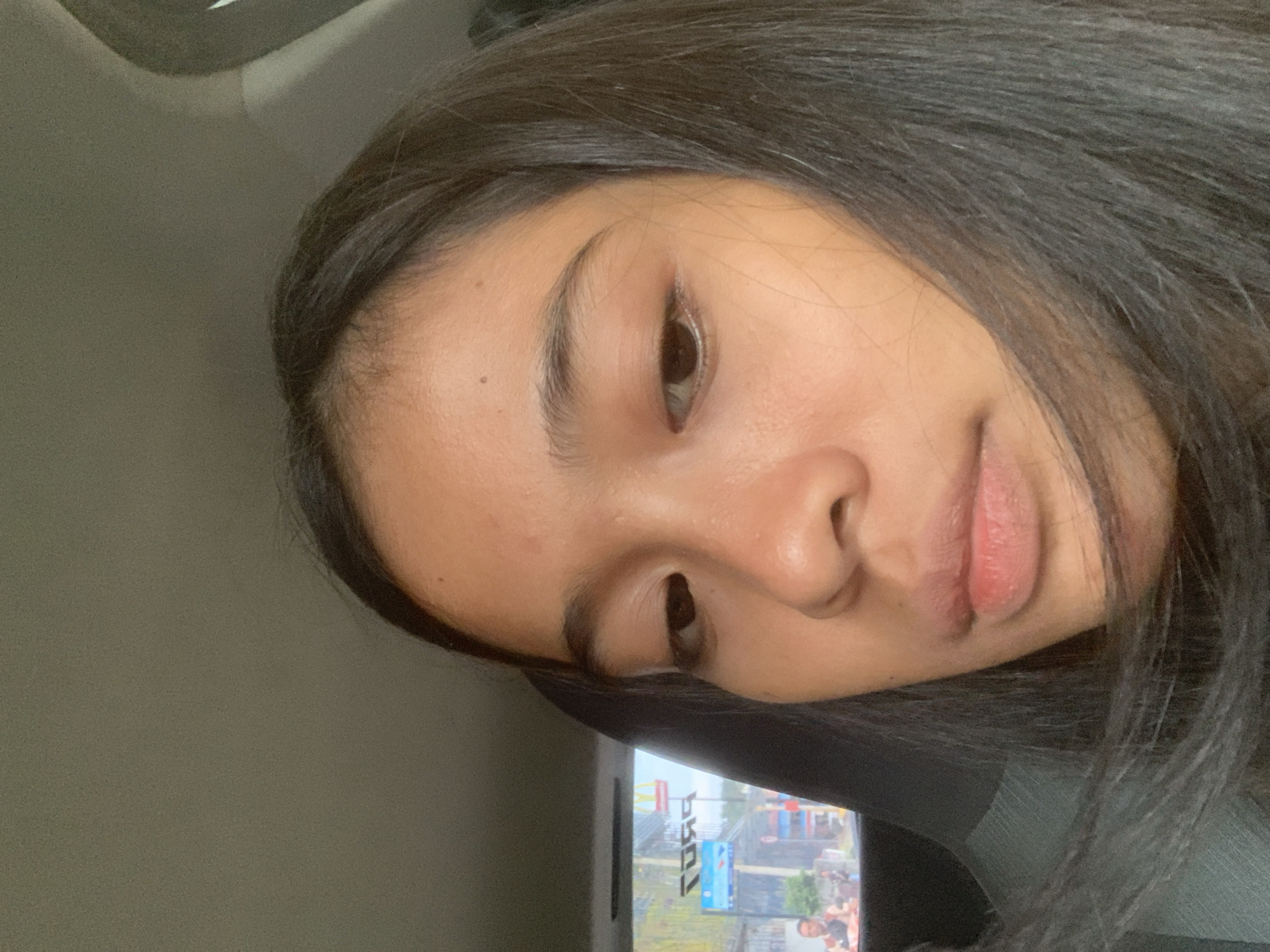 A close-up photo shows a young woman with dark hair and medium brown skin tone. She has dark eyebrows and eyes, and her expression is neutral. She is likely sitting in a vehicle, with a car interior visible in the background. The window of the car shows the outside view of Samarinda, Indonesia, with an advertisement of "TORQ" written on it, and buildings with flags displayed. The background is slightly blurred.