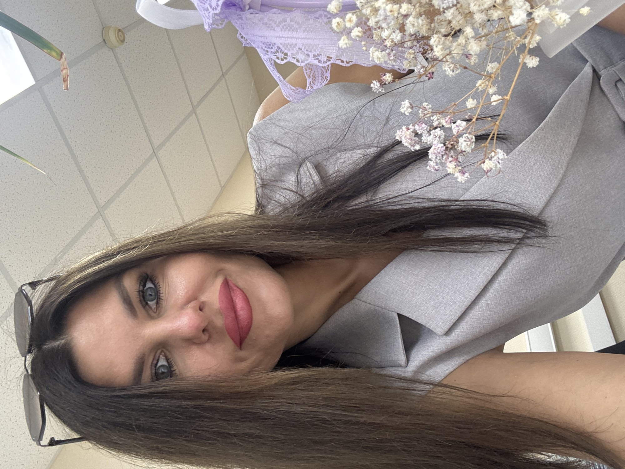 The image is a close-up, selfie-style shot of a woman with long, dark brown hair and blue eyes, wearing a light gray sleeveless blazer. She has on pink lipstick and sunglasses perched on her head. In the foreground, a bouquet of small white and pink dried flowers is visible. In the background, a white tiled ceiling with fluorescent lighting is apparent. The overall setting appears to be an indoor environment, possibly an office or a similar professional space, given the ceiling tiles and lighting. There is no identifiable context within the image to indicate a specific location in Chaikovsky, Russia.