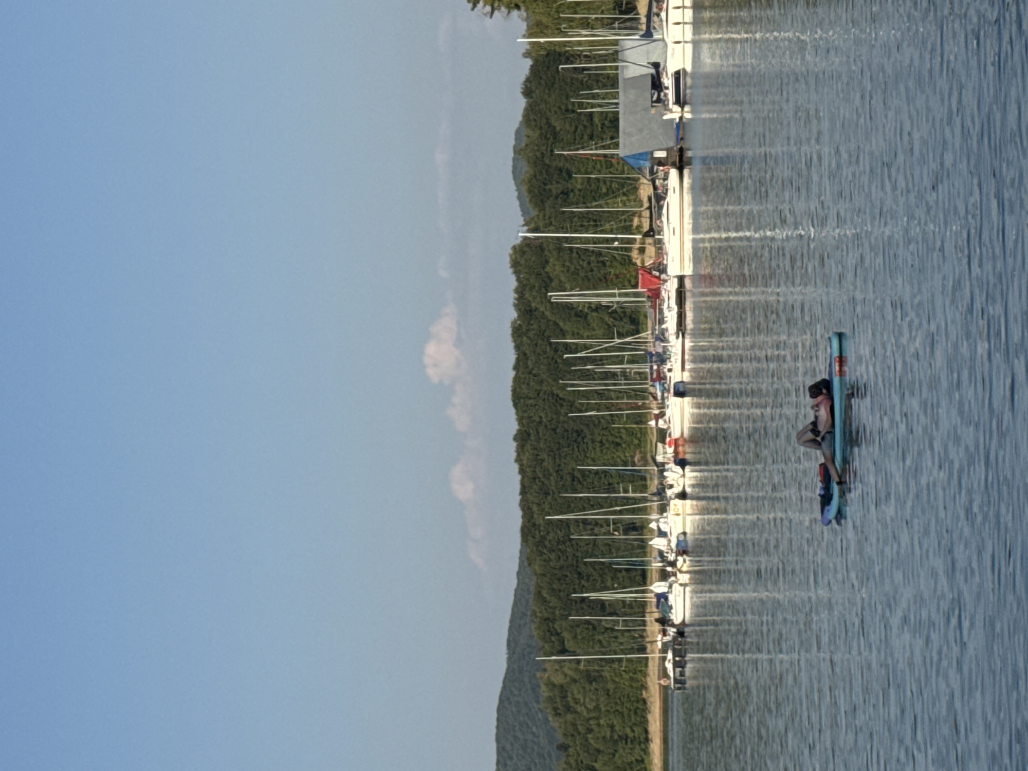 In Polańczyk, Poland, a man lies on a stand-up paddleboard in a body of water. The paddleboard is blue and purple. The man is wearing dark shorts and is lying on his back. In the background, a marina is visible with numerous sailboats docked. The boats have white hulls and tall masts, some with sails furled. Beyond the marina, a dense green forest covers rolling hills under a clear blue sky with a few wispy white clouds. The water is calm and reflects the sky and the boats.