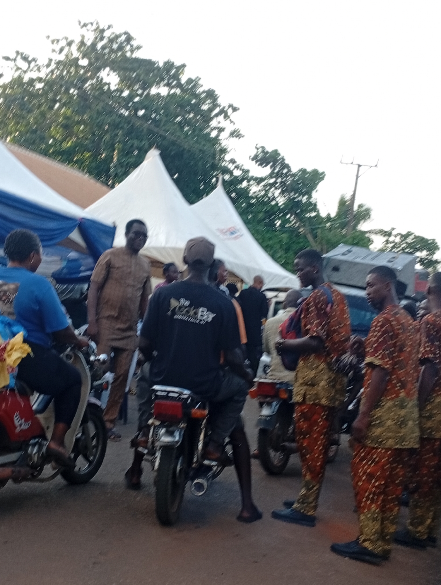 The image depicts an outdoor gathering in Nsugpe, Nigeria, under a bright sky with some trees visible. In the foreground, two motorcycles are parked, one red and one darker colored. Several individuals are present, most of whom are dressed in traditional Nigerian attire.
On the left, a woman wearing a blue t-shirt and dark pants is seated on the red motorcycle, looking towards the center. A man in a brown, patterned traditional outfit is standing between the motorcycles, facing the viewer and smiling. To his right, a person wearing a dark t-shirt with "The Rock Bay" and some text in a yellow graphic is also near the motorcycles.
Further to the right, three men are standing together, all wearing matching orange and gold patterned traditional outfits. They are looking in different directions. Behind them, a large white tent with blue draping is set up, suggesting an event or market. A utility pole is visible in the background on the right. The ground appears to be paved, likely a road or open area within the city. The overall impression is of a community event or a busy marketplace.