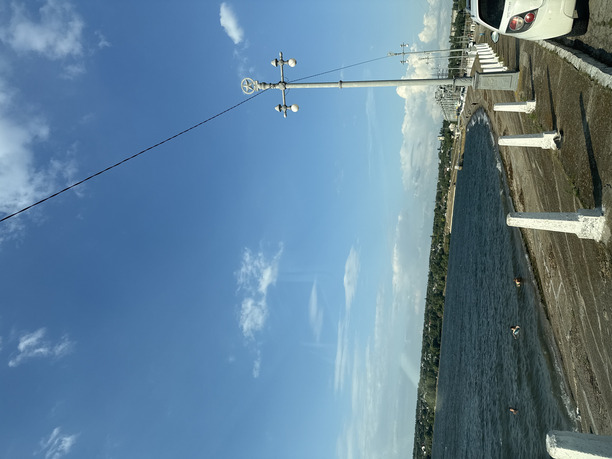 The image depicts a waterfront scene with a wide body of dark blue water on the left and a paved walkway with decorative white bollards and lampposts on the right. The sky is bright blue with scattered white clouds. In the water, three individuals are visible, appearing to be swimming or wading. The shoreline consists of a sloped embankment leading to the water. Further inland, a distant tree-lined town is visible, and beyond that, a large industrial structure and a bridge can be seen. A white car is parked on the right side of the walkway. A thin black wire stretches diagonally across the upper portion of the image.