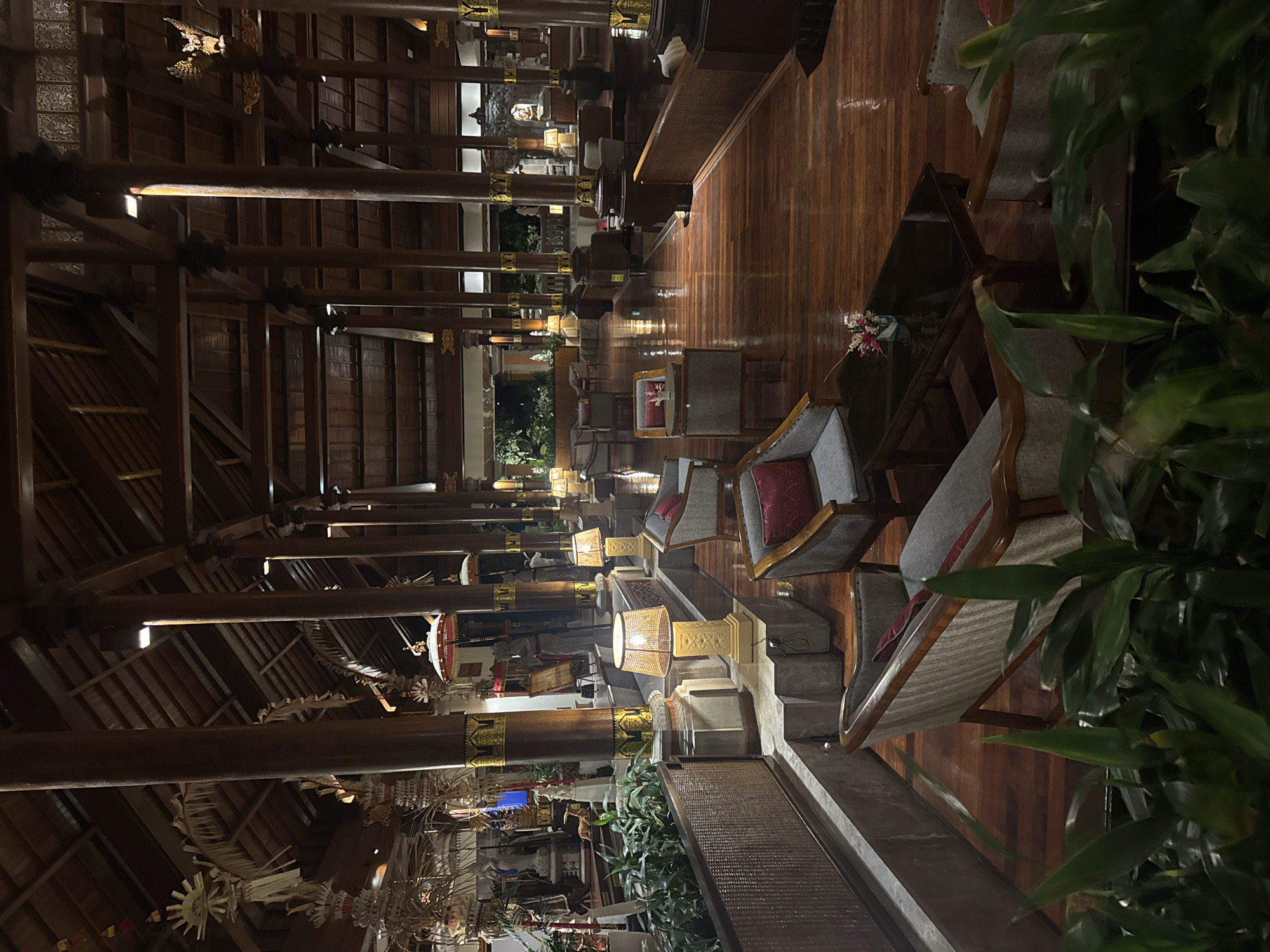 An interior view of a grand lobby in Kuta Selatan, Indonesia, features a high, wooden beamed ceiling supported by large, dark wooden pillars with ornate gold accents. The polished wooden floor reflects the warm glow of ambient lighting. Several seating areas with plush armchairs and sofas are strategically placed throughout the space, some with red accent pillows. Small, dark wood coffee tables accompany some of the seating arrangements. Decorative lamps with patterned shades illuminate the space, and lush green plants in woven planters add a touch of nature. In the background, traditional Balinese architectural elements and decorative items are visible. The overall impression is one of luxurious tranquility and cultural richness, typical of a high-end resort or hotel lobby.