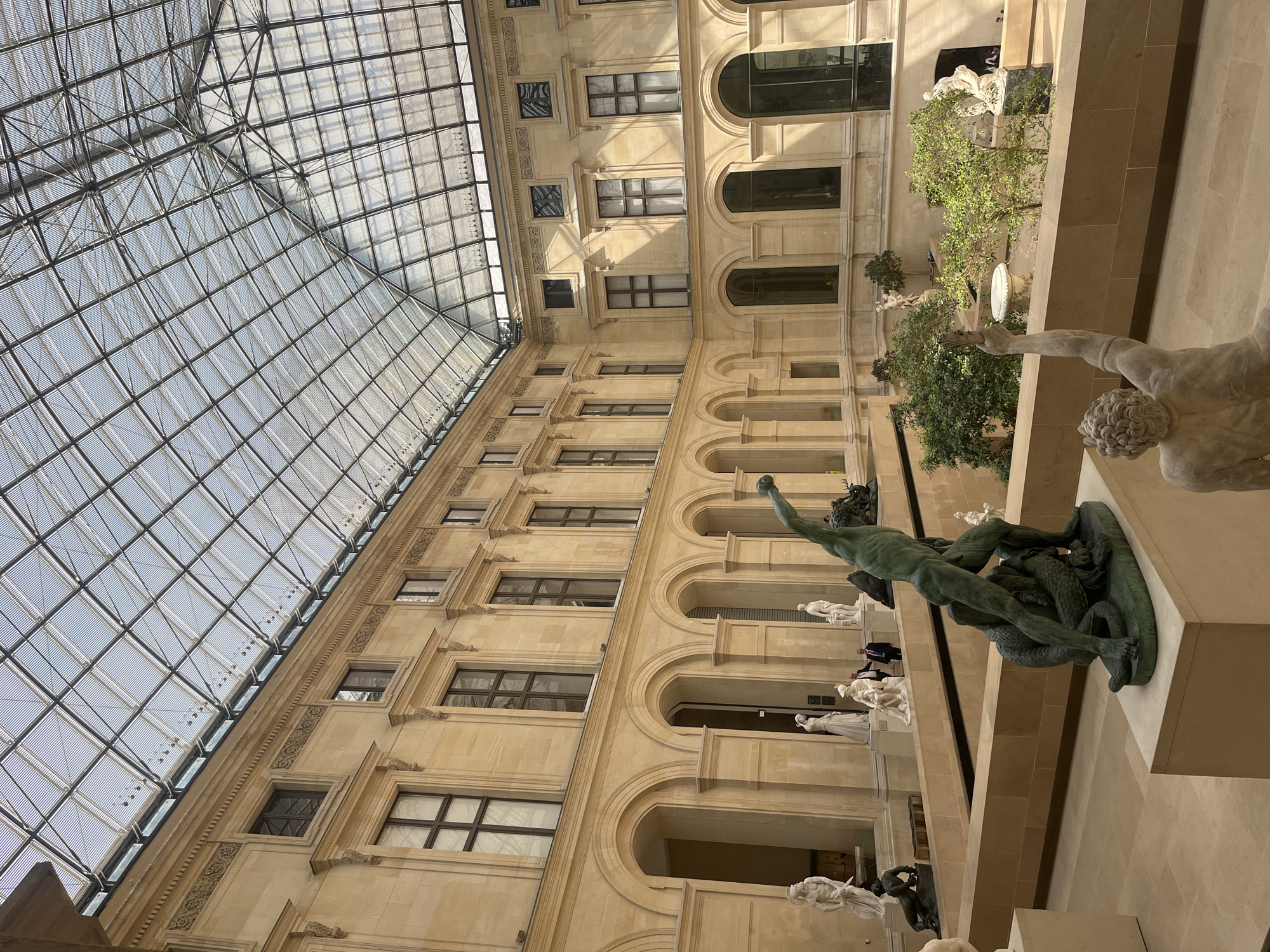 This image captures the magnificent interior of a grand museum or gallery, located in Paris, France, characteristic of the Louvre Museum. The scene is bathed in natural light filtering through an expansive, geometrically gridded glass ceiling that forms a large vaulted skylight.
Below the soaring glass roof, the architectural elements feature multi-tiered walls constructed from light-colored stone, adorned with rows of arched alcoves and rectangular windows. Intricate carvings and classical detailing embellish the cornices and sections between the levels, contributing to the historical and opulent atmosphere.
The space is populated with classical sculptures. Most notably, a prominent dark green (likely bronze) statue in the midground depicts a muscular male figure engaged in struggle with a serpent or sea monster. This is complemented by several white marble statues positioned on lower levels and within various alcoves, portraying nude or draped figures in classical poses. Small trees and bushes are placed in planters, adding touches of greenery throughout the indoor environment.
A few human figures, appearing as visitors or staff, are visible in the background, providing a sense of scale within the vast interior. The bright, diffused illumination suggests daytime. A partial and indistinct word, "VAI", is visible on a high wall in the background. The overall scene exudes a profound sense of cultural heritage and artistic grandeur.