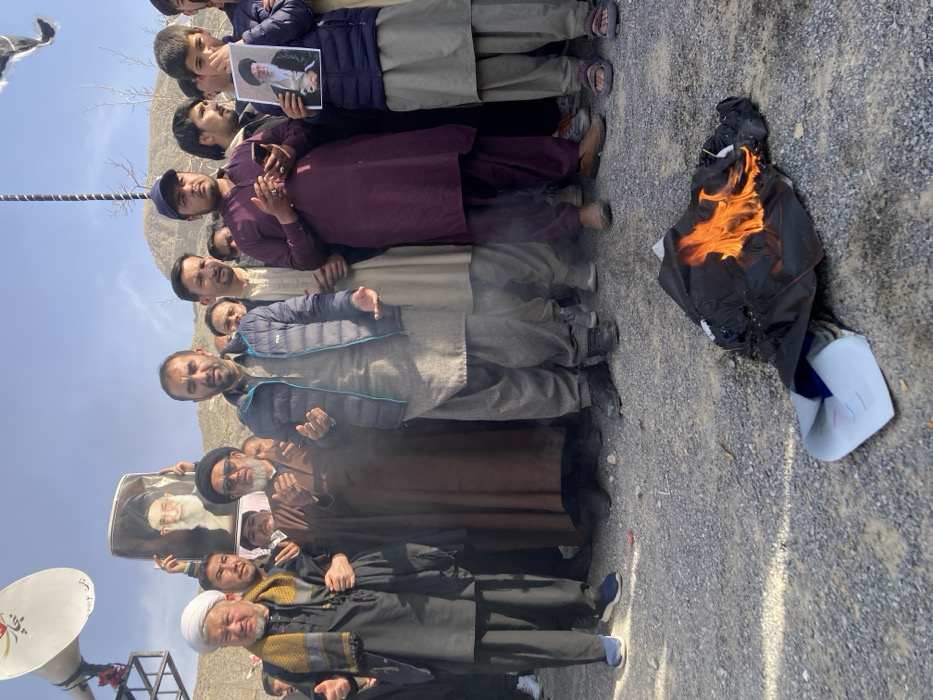 The scene is outdoors in Quetta City Tehsil, Pakistan, under a clear sky, with rocky mountains visible in the distant background. A group of approximately 15 to 20 males, including adults and youths, are present. They are primarily dressed in shalwar kameez; some wear turbans or caps. In the immediate foreground, a black garment, possibly a jacket or shirt, is actively burning on a light-colored gravel and stone ground, producing bright orange flames and grey smoke. Many individuals in the crowd have their hands raised to chest height, palms open and facing upwards. On the left side of the group, a man holds a large, framed portrait of an elderly man with a white beard and a black turban. On the right side, a young boy holds a smaller, unframed portrait of an elderly man with a white beard and a black turban, appearing distinct from the larger portrait. A prominent white megaphone is mounted on a metal support structure in the upper left background. One man is observed holding a smartphone in his hands.