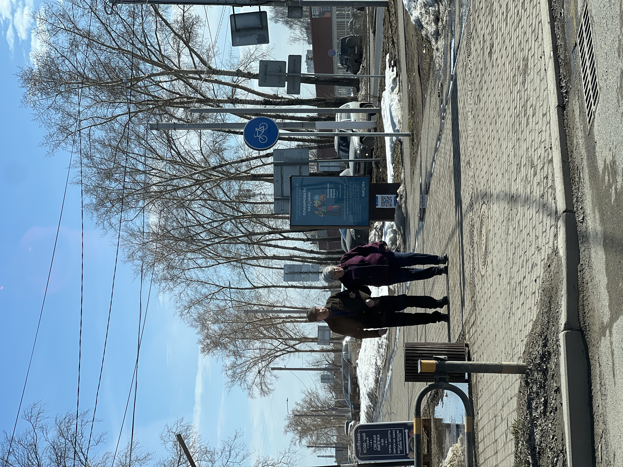 The image depicts an urban street scene in Chaikovsky, Russia, under a clear blue sky with sparse clouds, indicating bright daylight. The ground shows evidence of melting snow, suggesting early spring or late winter. Two elderly individuals, a man and a woman, walk arm-in-arm from left to right on a paved sidewalk. The man, positioned on the left, wears a dark cap, a dark jacket, and dark trousers. The woman, on his right, wears a grey beanie, a dark purple jacket, and dark trousers.
In the immediate foreground to the left, a black A-frame sign with white Russian text advertises a "Bakery - Coffee-Tea to go," "Fresh Pastries," "Bread," "Cakes + Desserts," and "Pies to order." Nearby, a metallic bicycle rack and a dark brown cylindrical trash receptacle are present on the sidewalk. Patches of melting snow and mud are visible along the sidewalk's edge and in unpaved areas.
Further along the sidewalk, a large vertical billboard displays an advertisement in Russian related to financial planning. Adjacent to it, a blue circular traffic sign with a white bicycle symbol indicates a bicycle path or route. Multiple utility poles, streetlights, and other traffic signs are scattered throughout the midground. The street is lined with bare deciduous trees, possibly birches, whose branches are devoid of leaves. Several parked vehicles, including SUVs and sedans, are visible on the street and in parking areas beyond the sidewalk. Overhead power lines span across the upper portion of the frame.